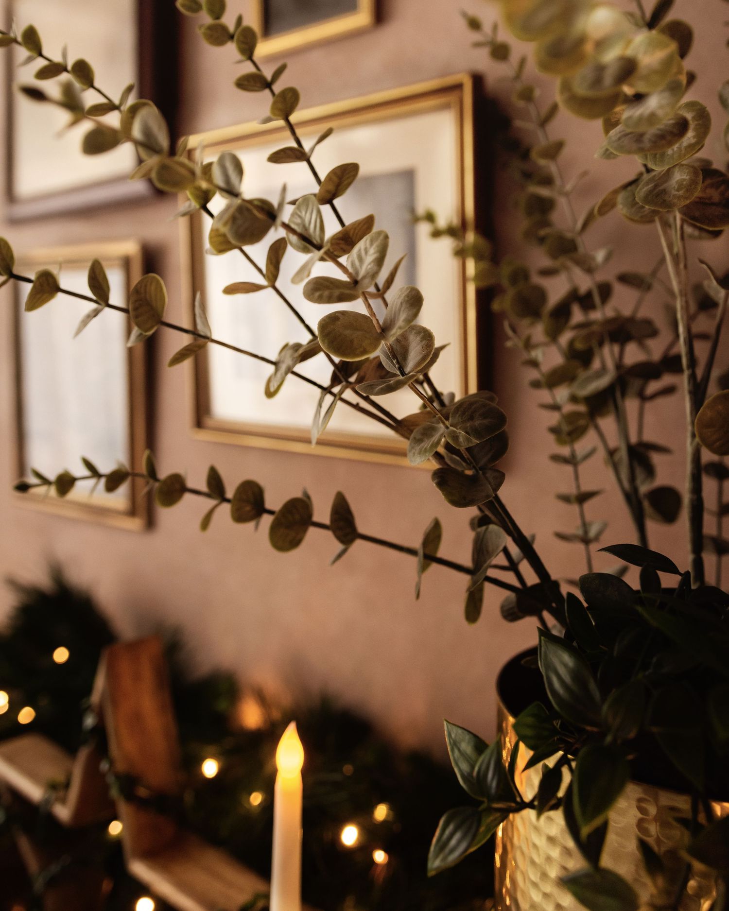 Cozy Christmas decor with eucalyptus garland, glowing candles, fairy lights, and gold-framed art on a warm-toned wall.