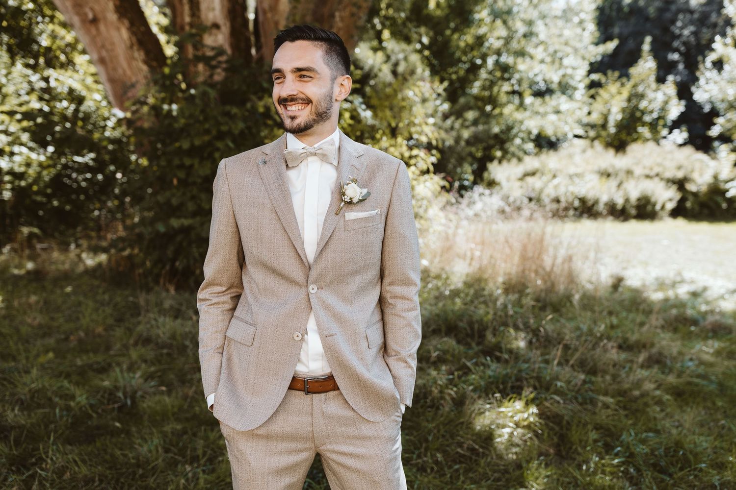 A person in a light beige suit stands outdoors in dappled sunlight among trees and tall grass.