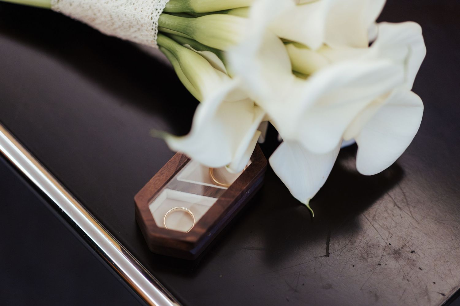 White calla lily flowers rest beside elegant wooden ring boxes on a dark textured surface.