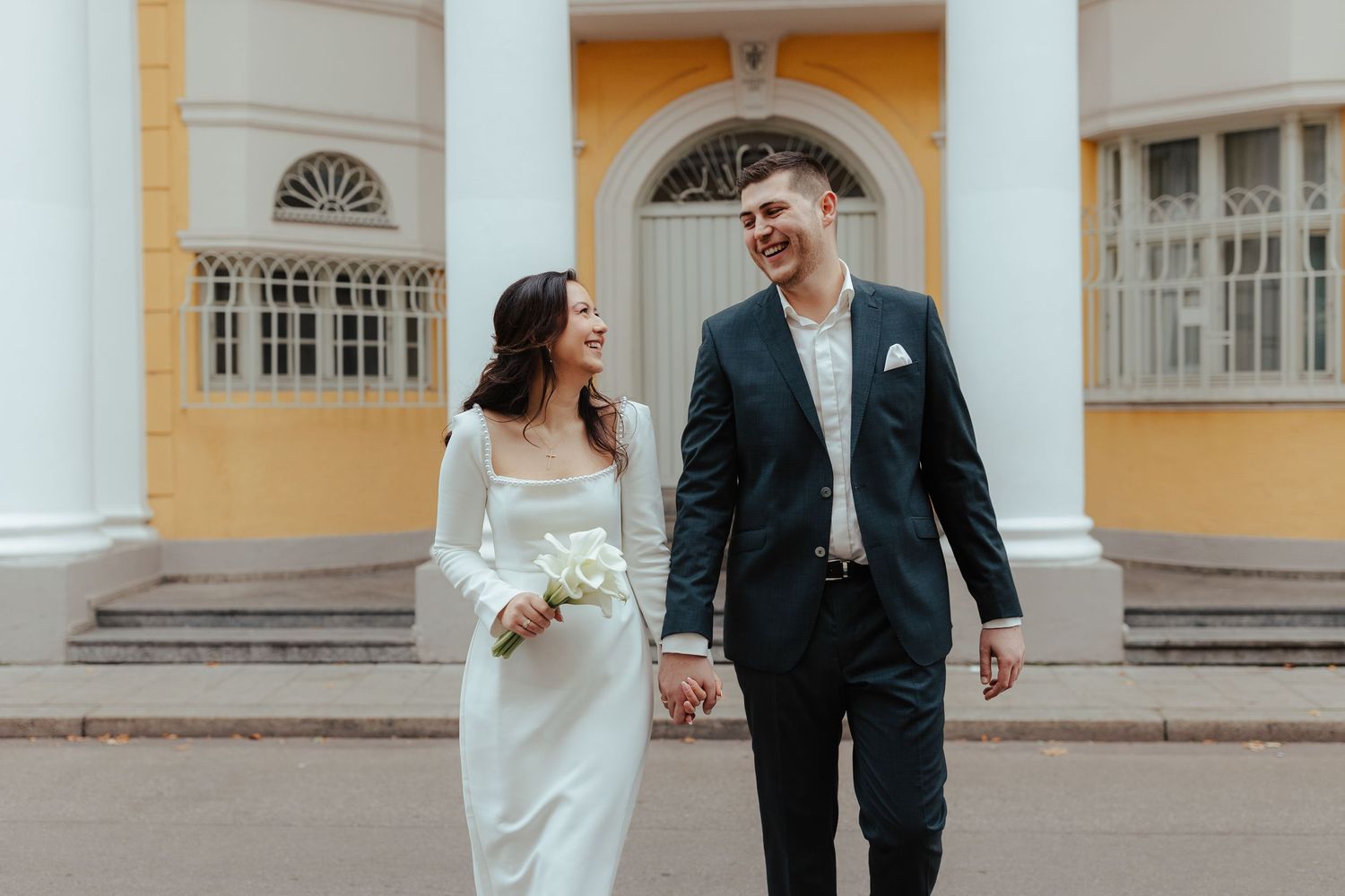 Couple in wedding attire walking and laughing together in front of a grand yellow building with arched windows.