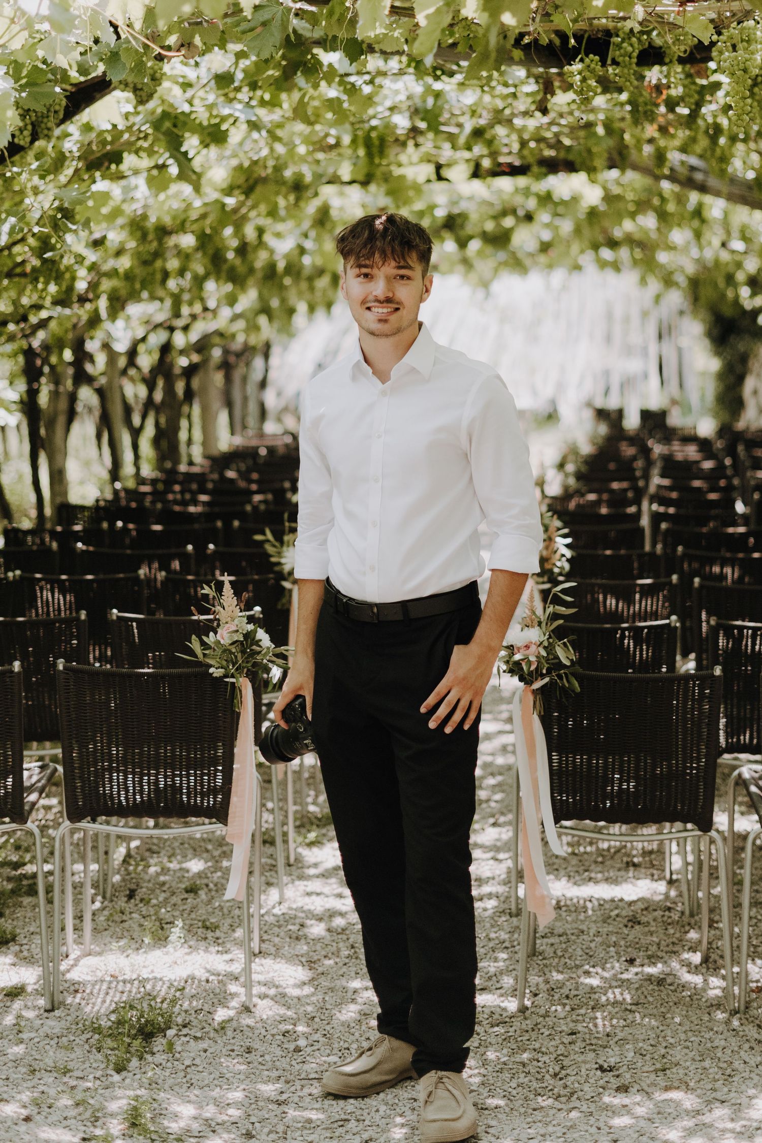 A person in a white shirt and black pants stands in a garden setting with white blossoms scattered on the ground.