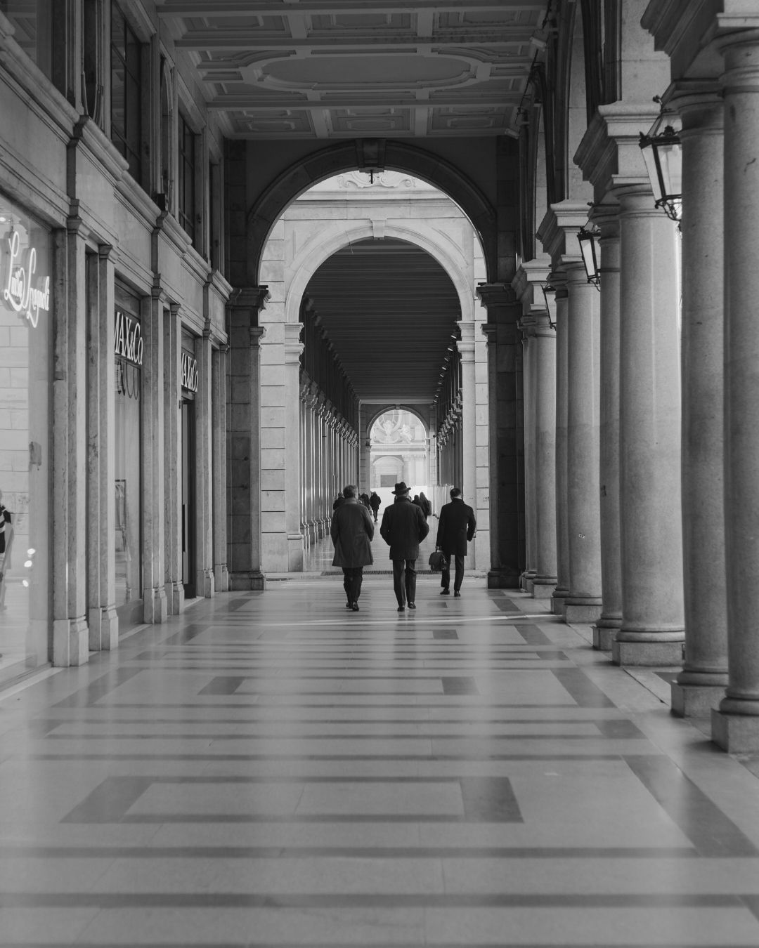 People walk through a historic covered colonnade with arched columns and repeating architectural patterns in black and white.