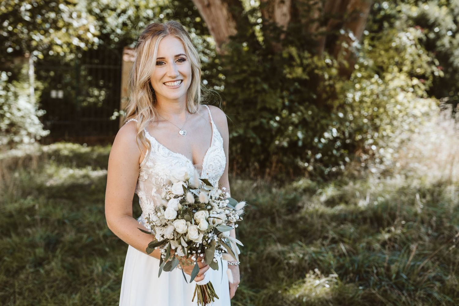 A bride in a white lace dress poses with a romantic bouquet of white and blush flowers in a sunlit garden setting.