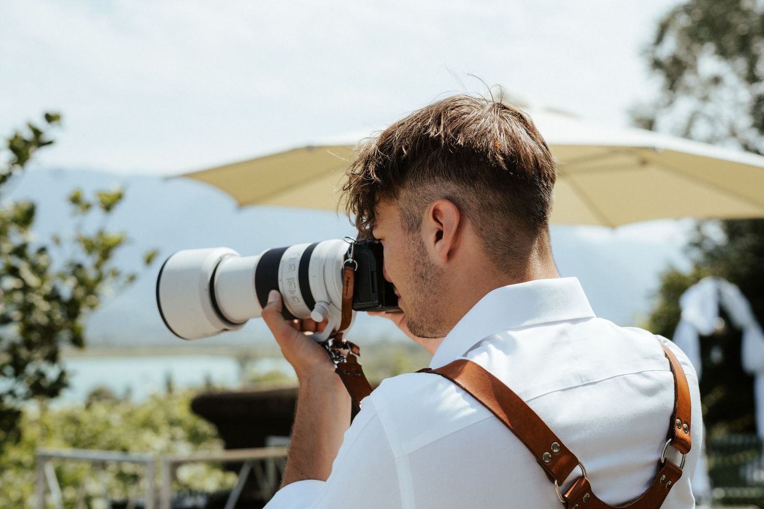 A photographer wearing a white shirt uses a telephoto lens outdoors on a sunny day.
