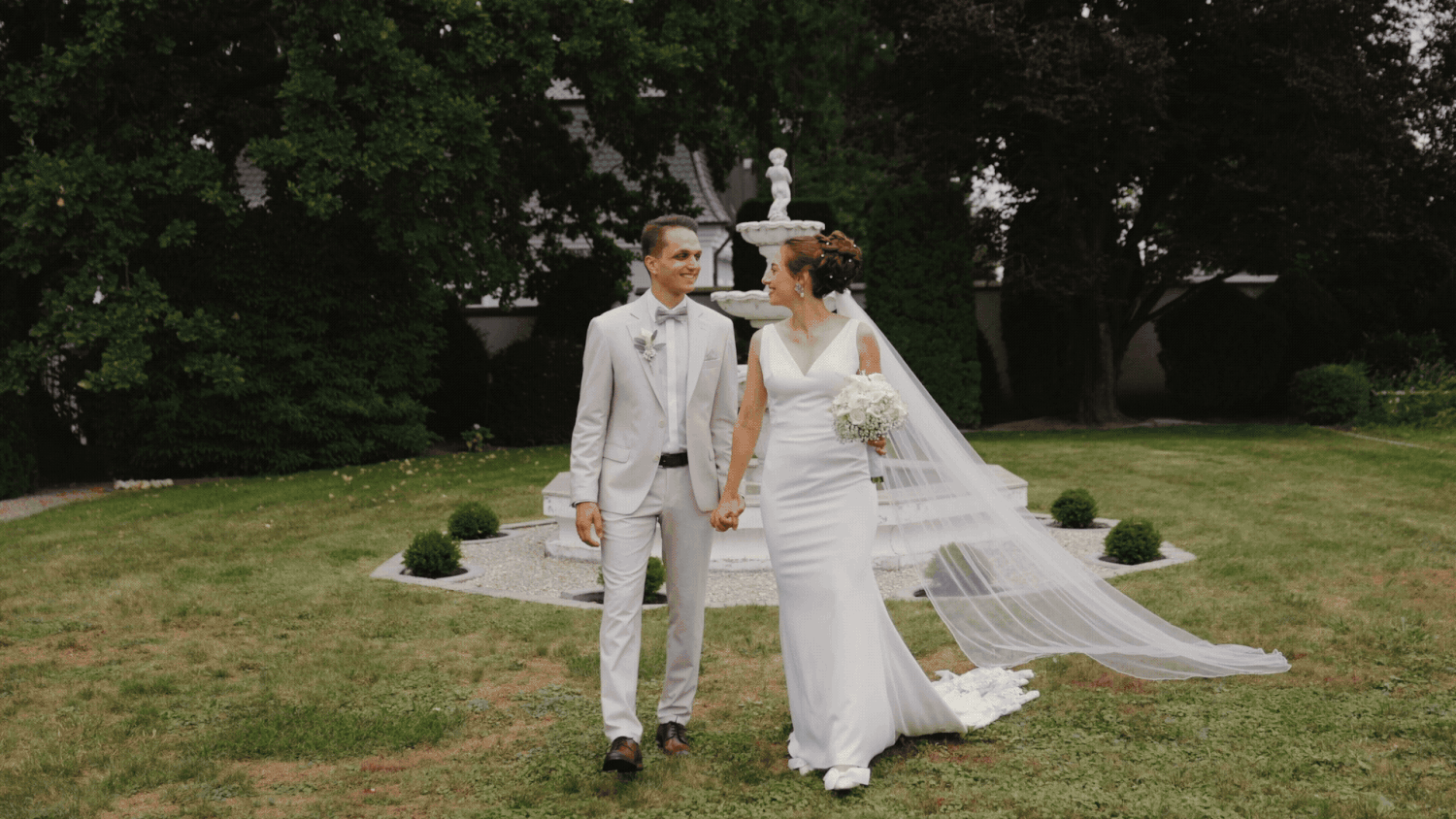 A couple in elegant white attire walks together on a grassy lawn during their wedding celebration.