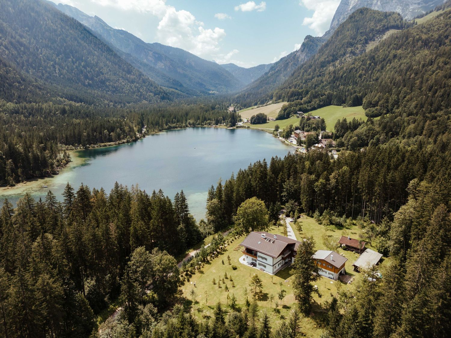 Panoramic view of an alpine lake surrounded by mountains, forests, and small buildings nestled in green meadows.