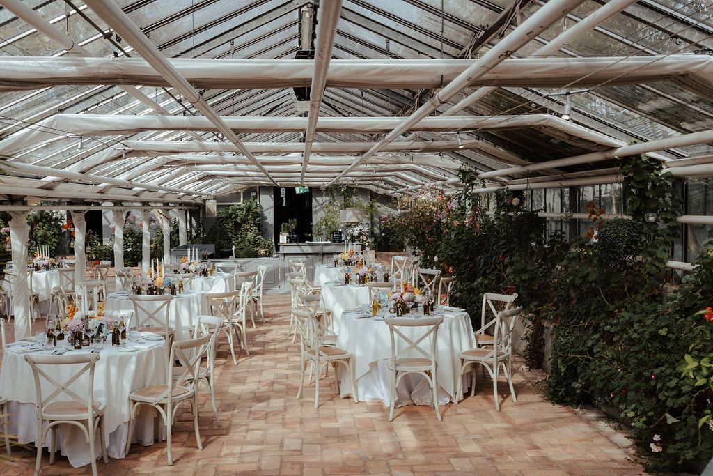 Elegant greenhouse wedding venue with white chairs, draped fabric, and natural light streaming through glass panels.