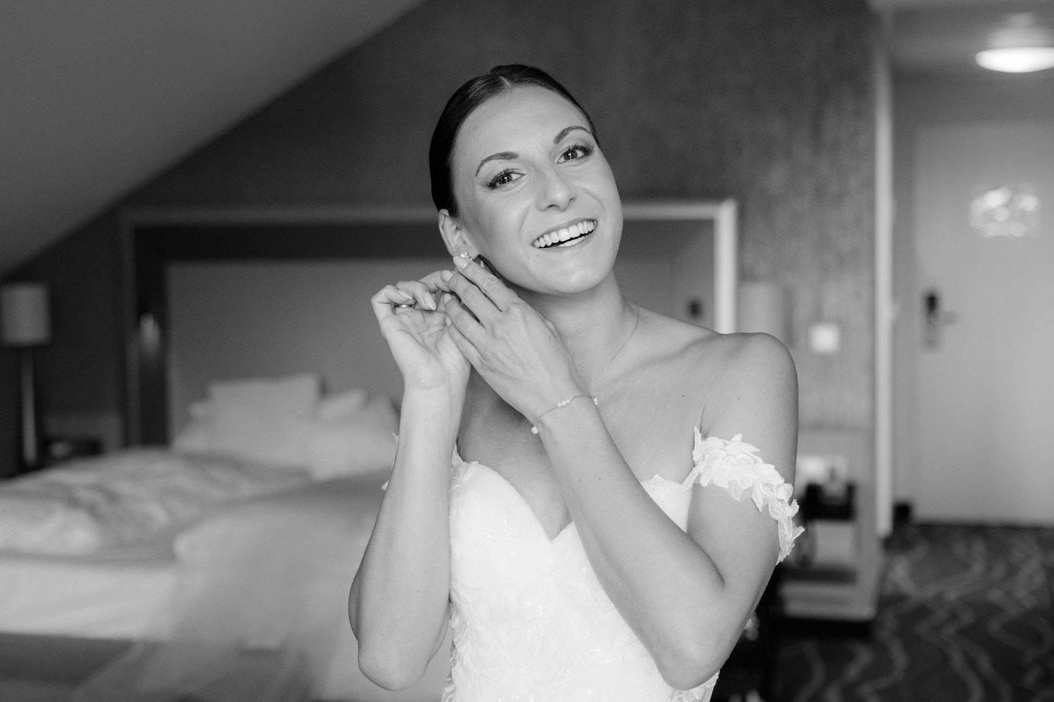 Black and white photo of a bride smiling while putting on earrings in a hotel room.