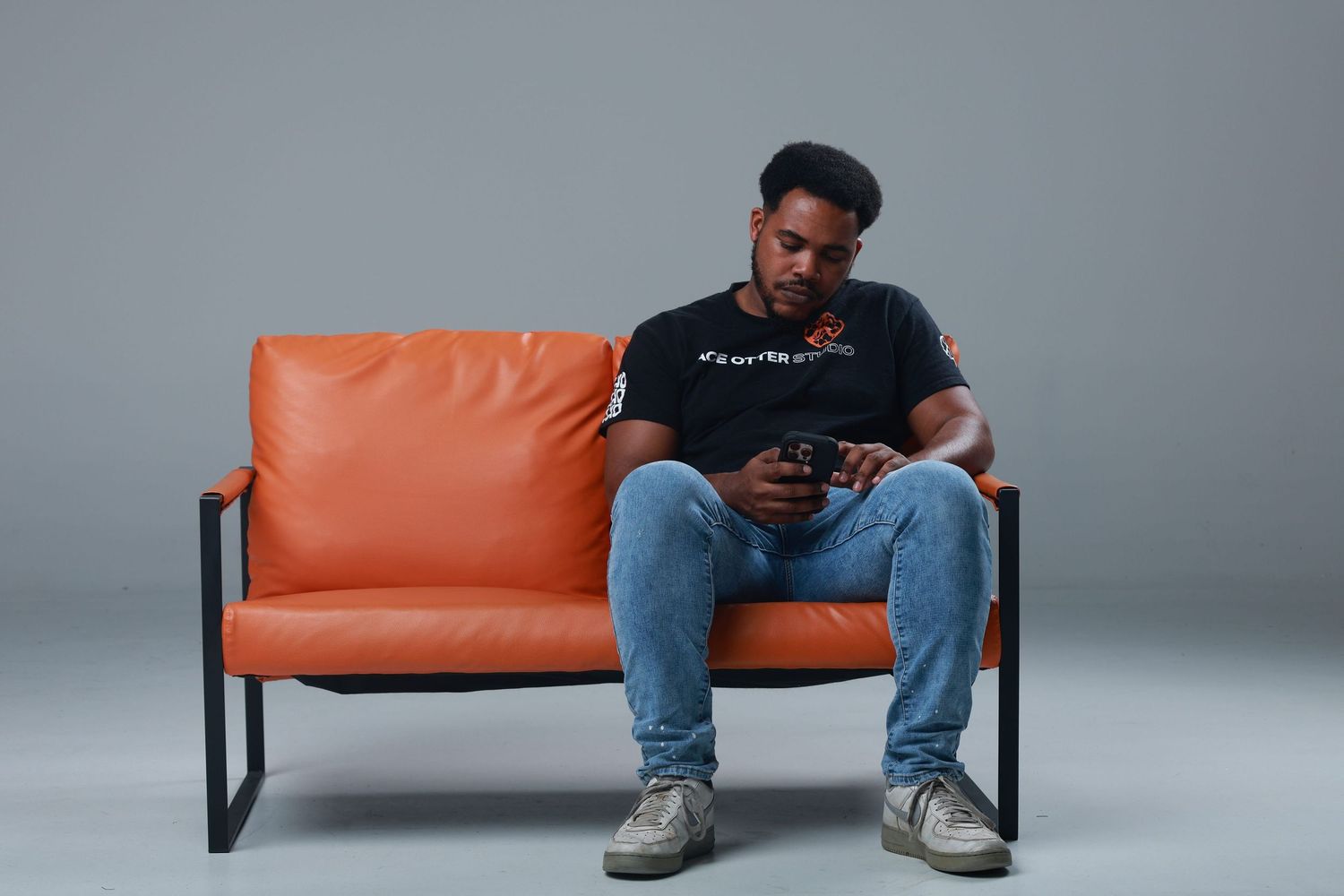 Man in black t-shirt and jeans sitting on orange leather sofa using smartphone against gray background.