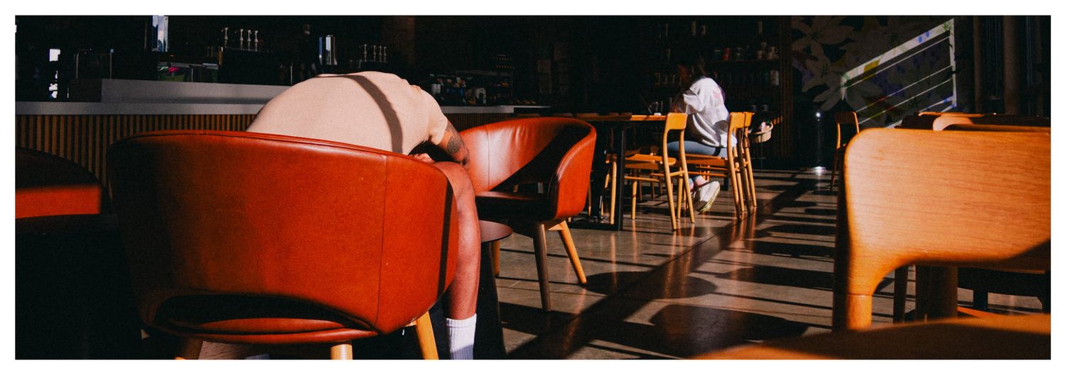 Person sitting alone in a dimly lit restaurant with red and wooden chairs on a checkered floor.