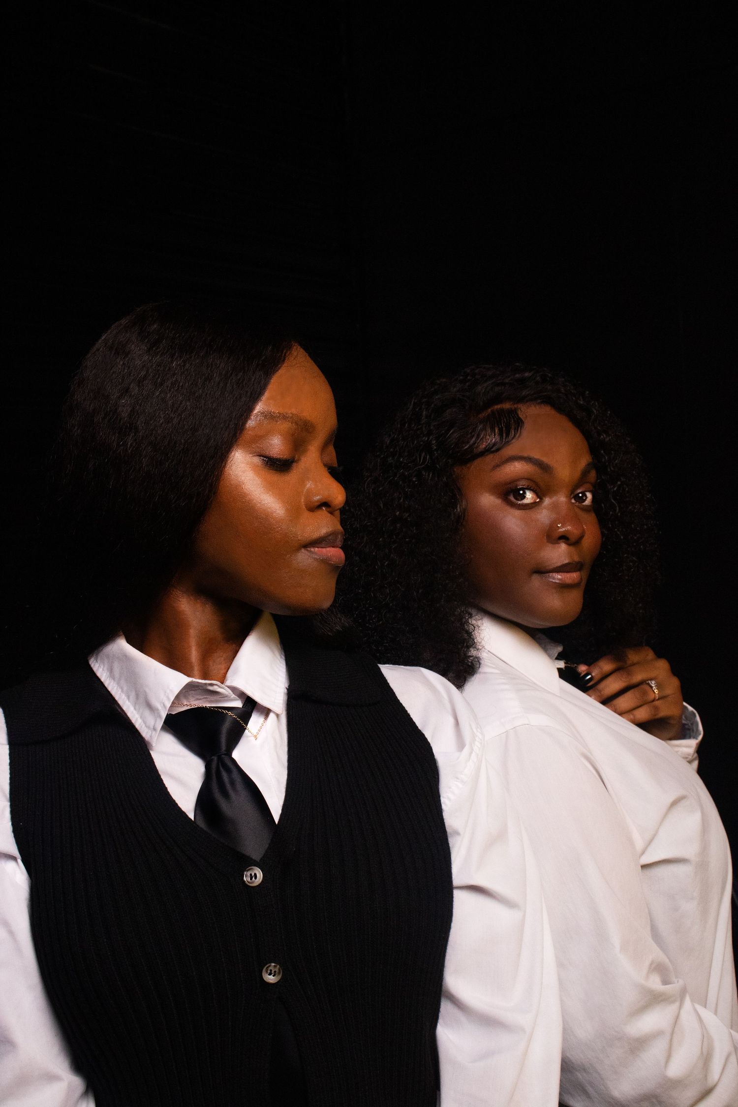 Two Black women in formal business attire posing together against dark black studio background.