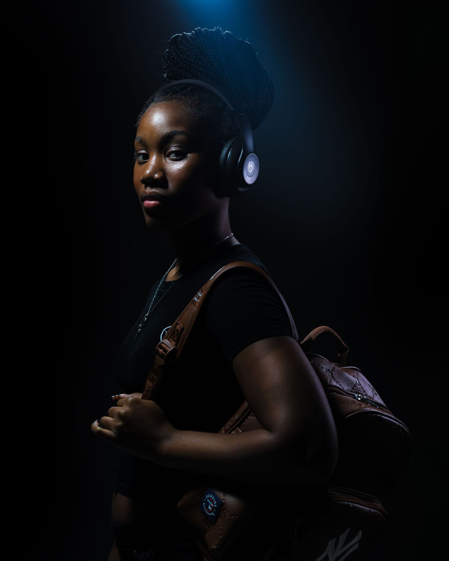 Young Black woman with braided updo and headphones, wearing black top and brown leather backpack, dramatic dark studio lighting.