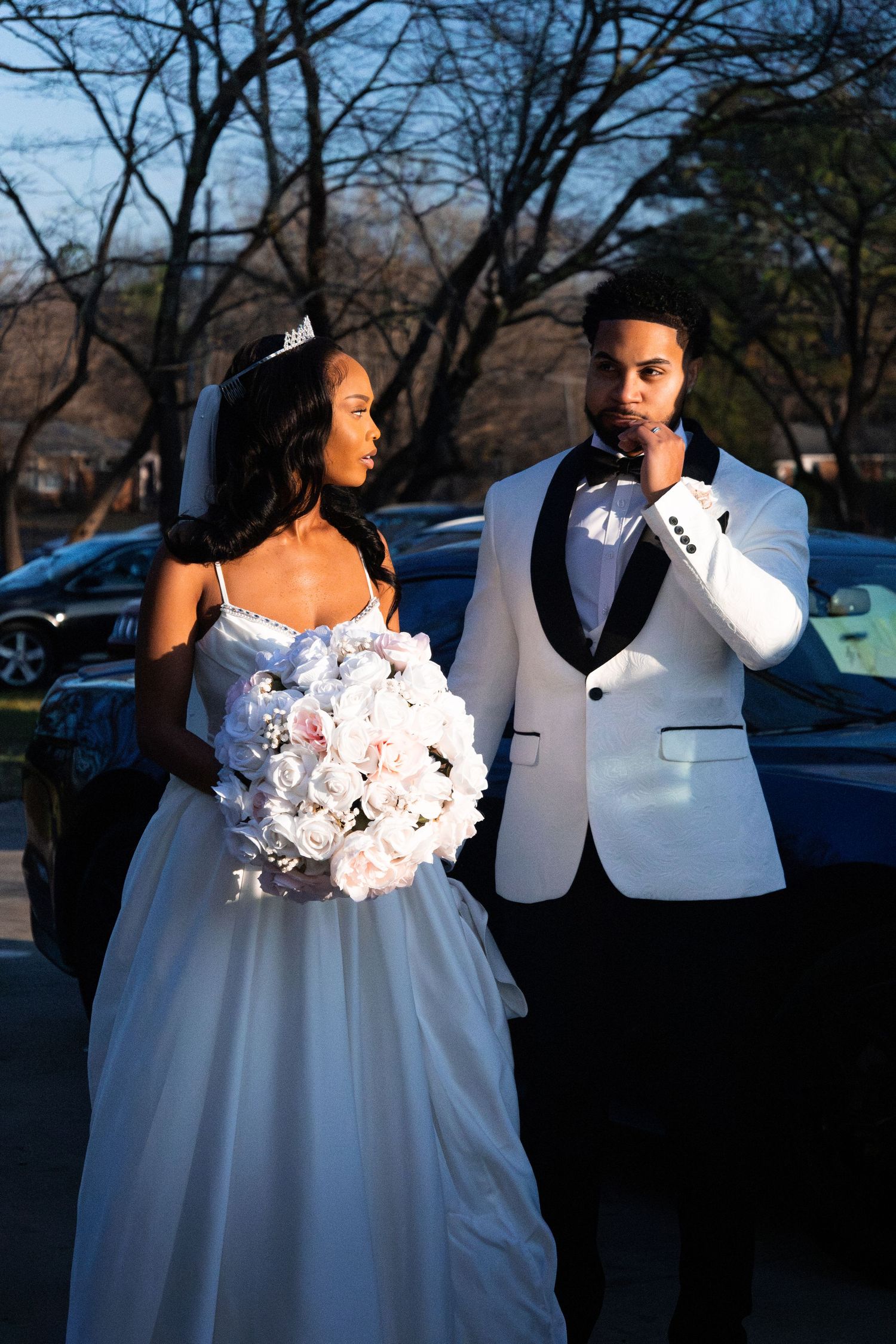 A bride in a white gown holding roses and a groom in a white tuxedo pose outdoors near trees at dusk.