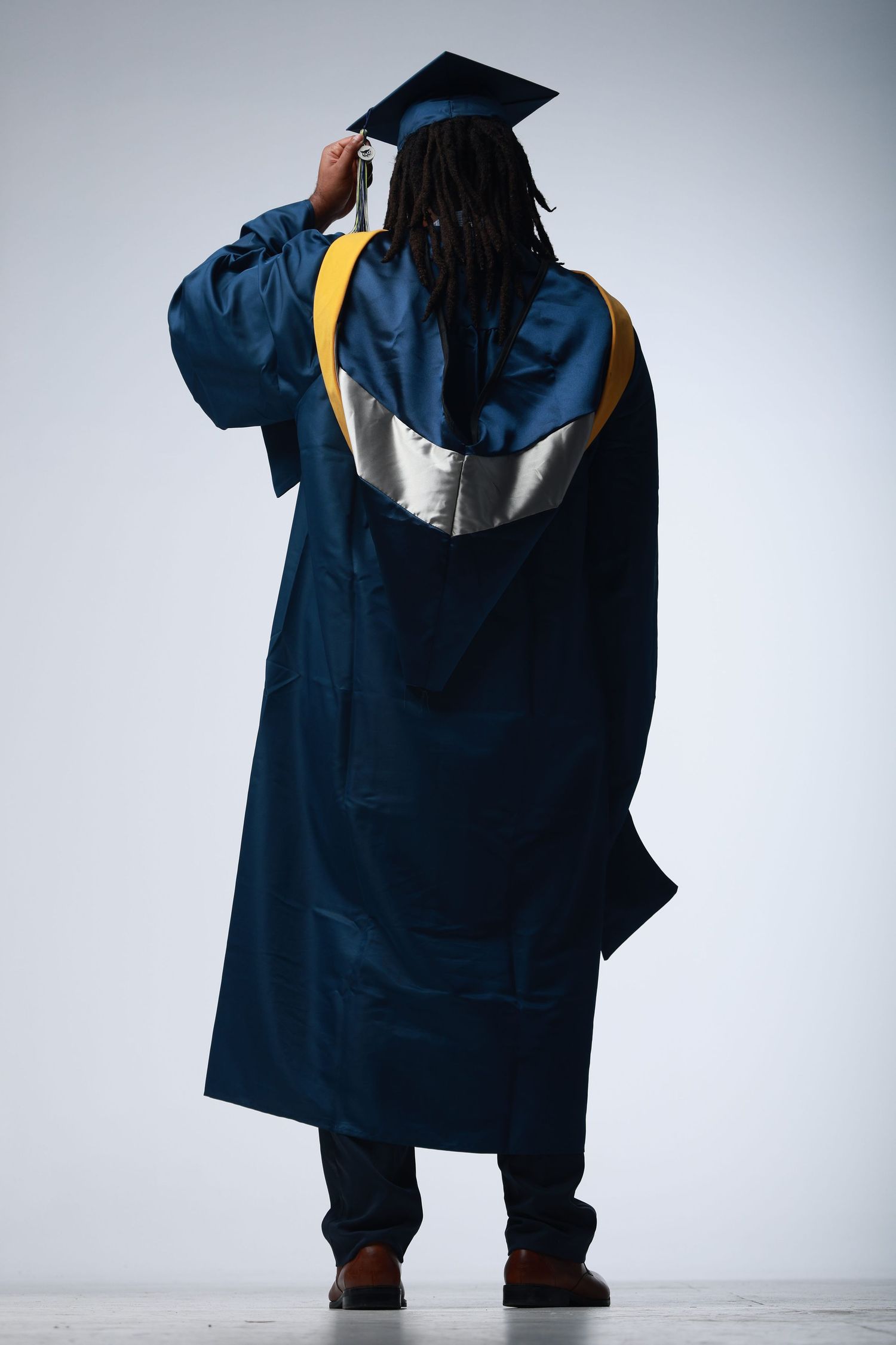 Graduate in navy blue cap and gown with gold hood, seen from behind on light gray background.