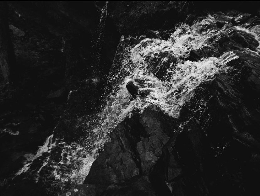 Black and white aerial view of rushing waterfall cascading over dark rocky terrain in dramatic contrast.