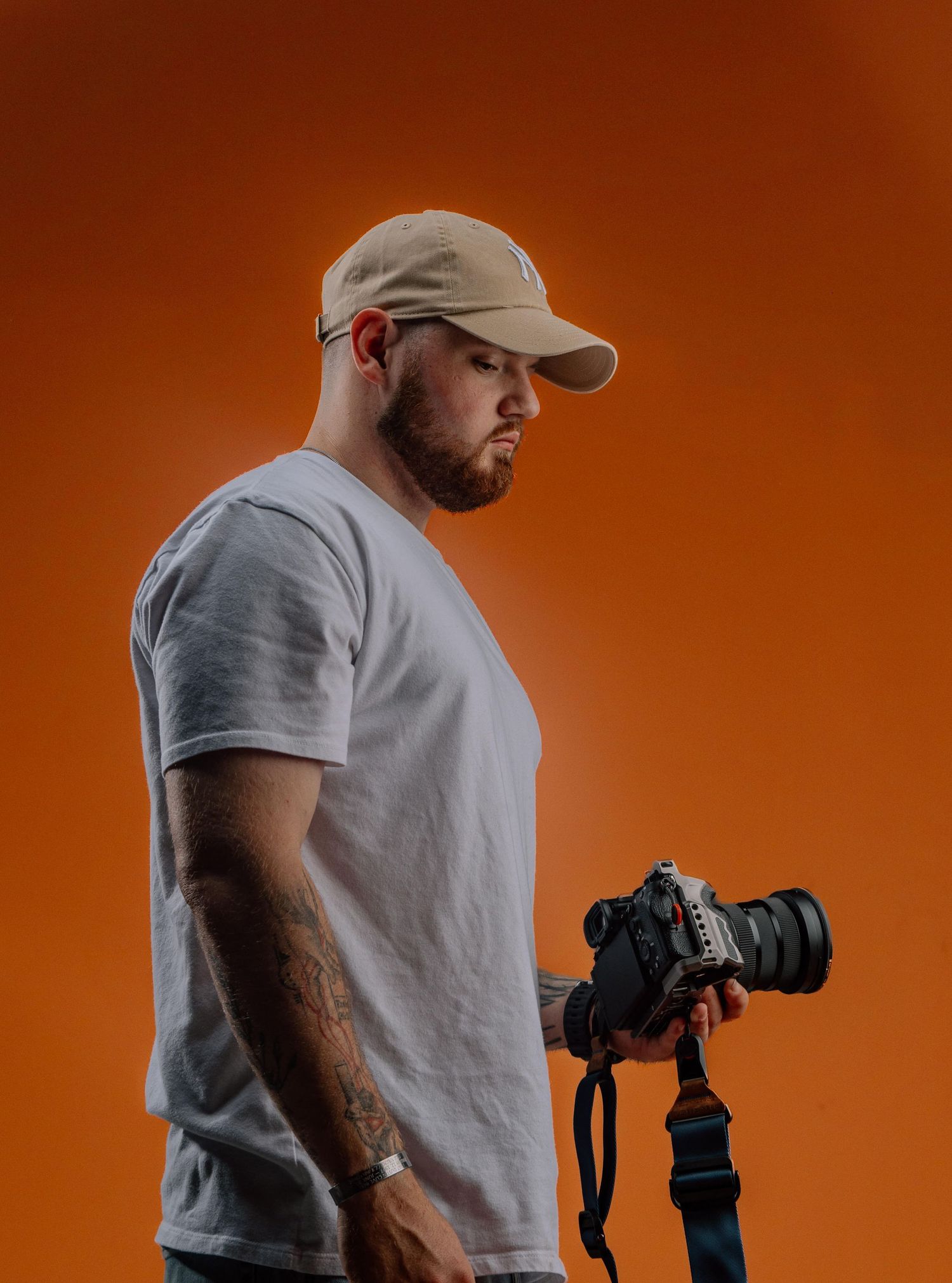 Tattooed male photographer in beige cap and white t-shirt holding DSLR camera against orange background.
