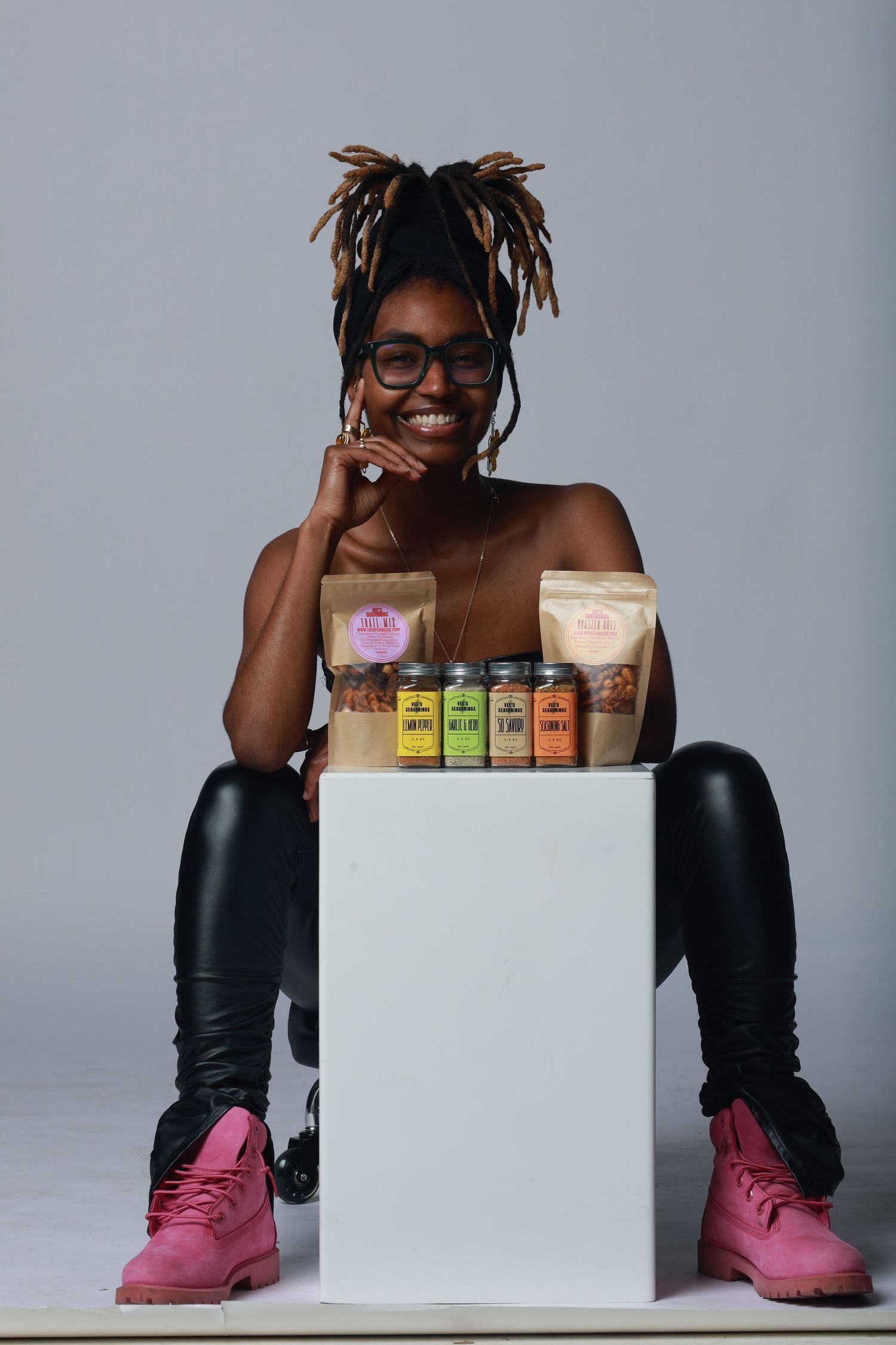 Smiling woman with dreadlocks in glasses and pink boots poses beside a white cube displaying spice jars and snack bags.