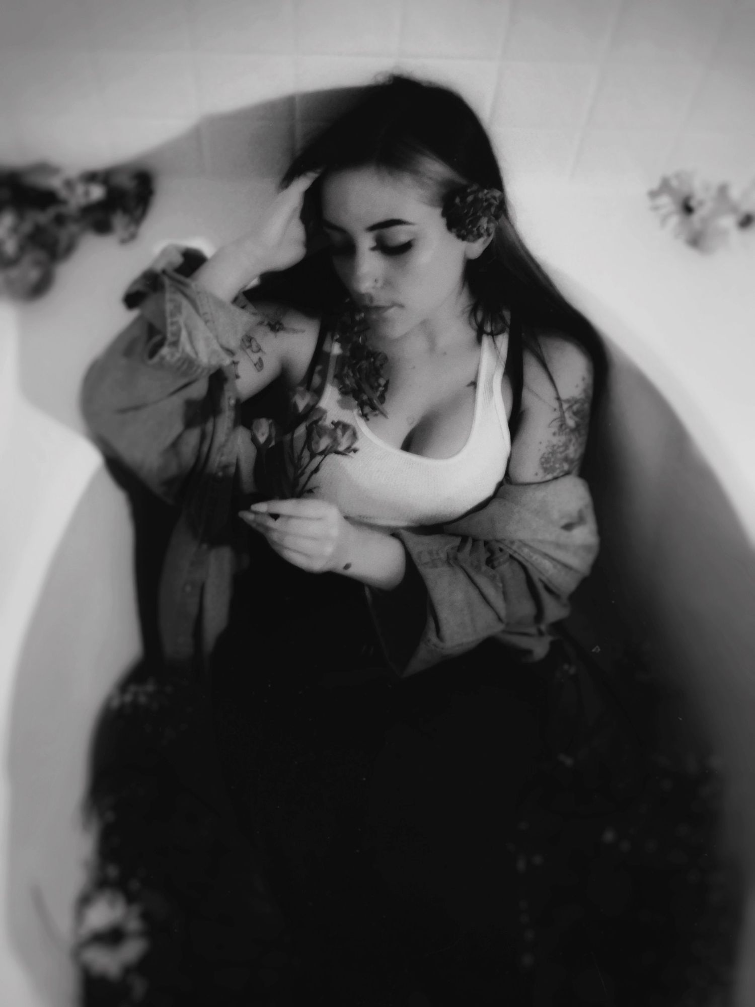 Black and white photo of tattooed woman sitting in bathtub with flowers.