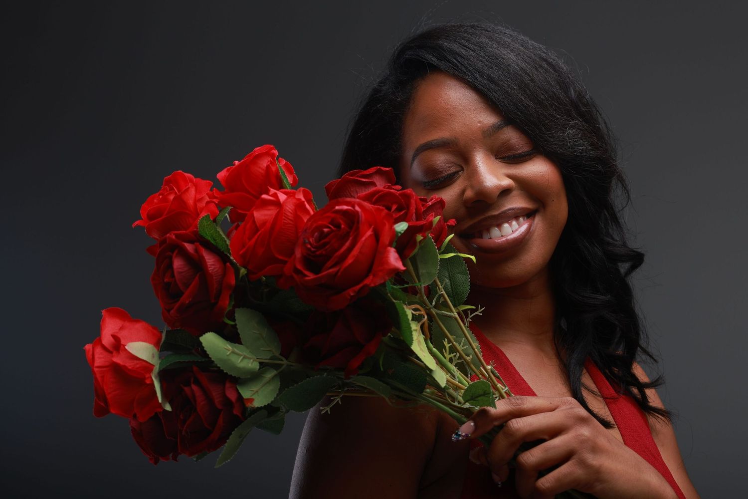 Smiling woman holding a bouquet of red roses, wearing a red top against a dark gray background.