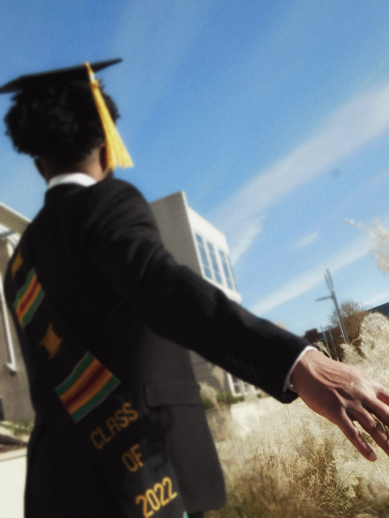 A 2022 graduate in cap and gown with colorful stole reaches out their hand outdoors on a sunny day.