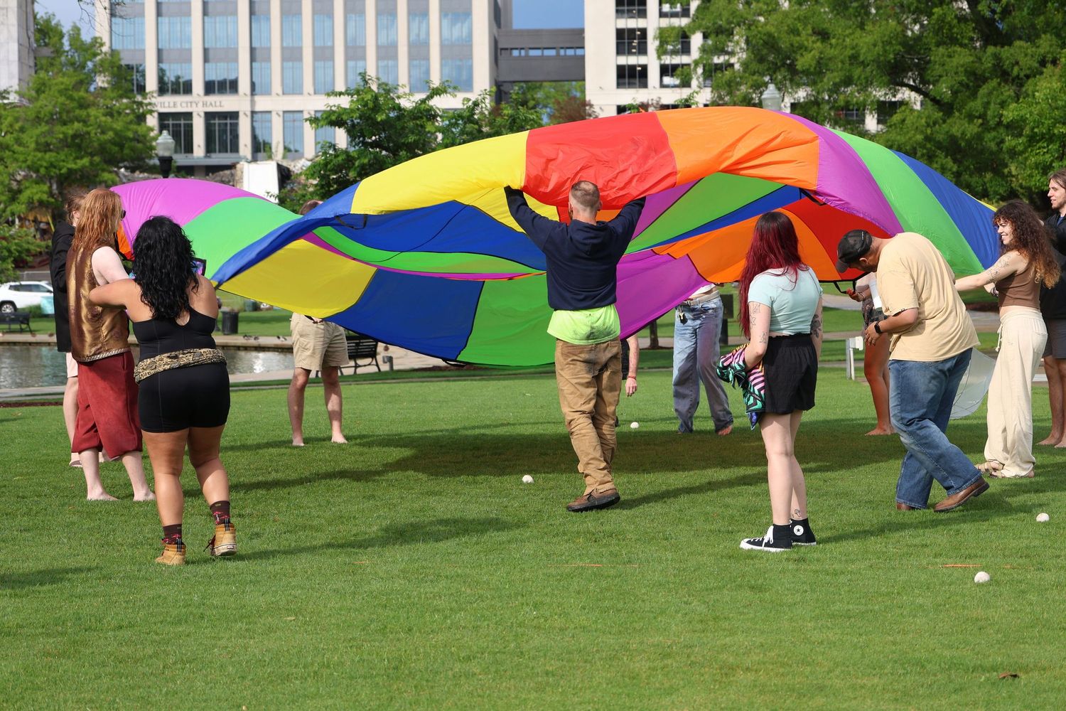 A group of people play with a large rainbow-colored parachute on a grassy lawn near office buildings.