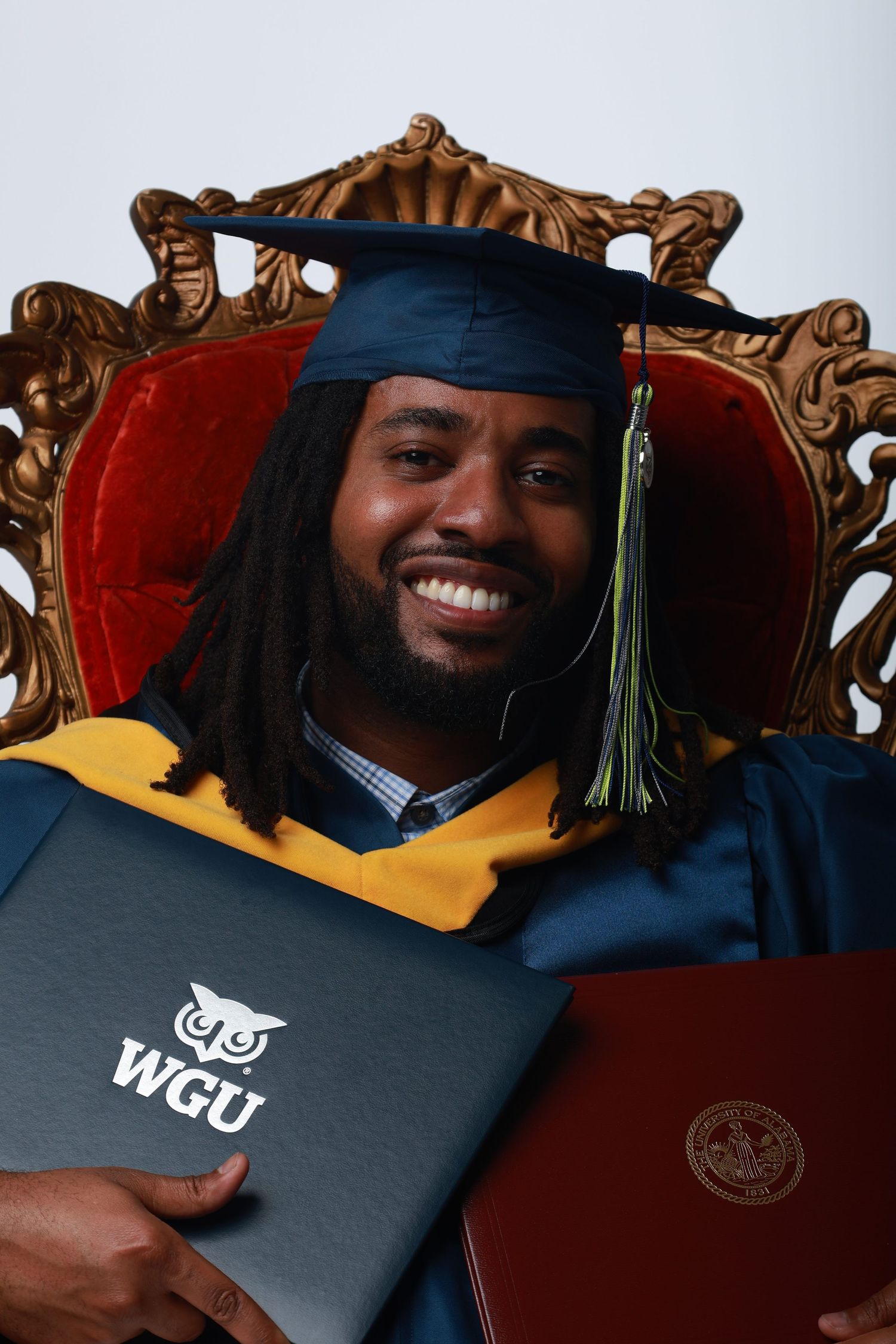 Smiling graduate in cap and gown holds two diplomas, including WGU degree, seated in ornate gold chair.