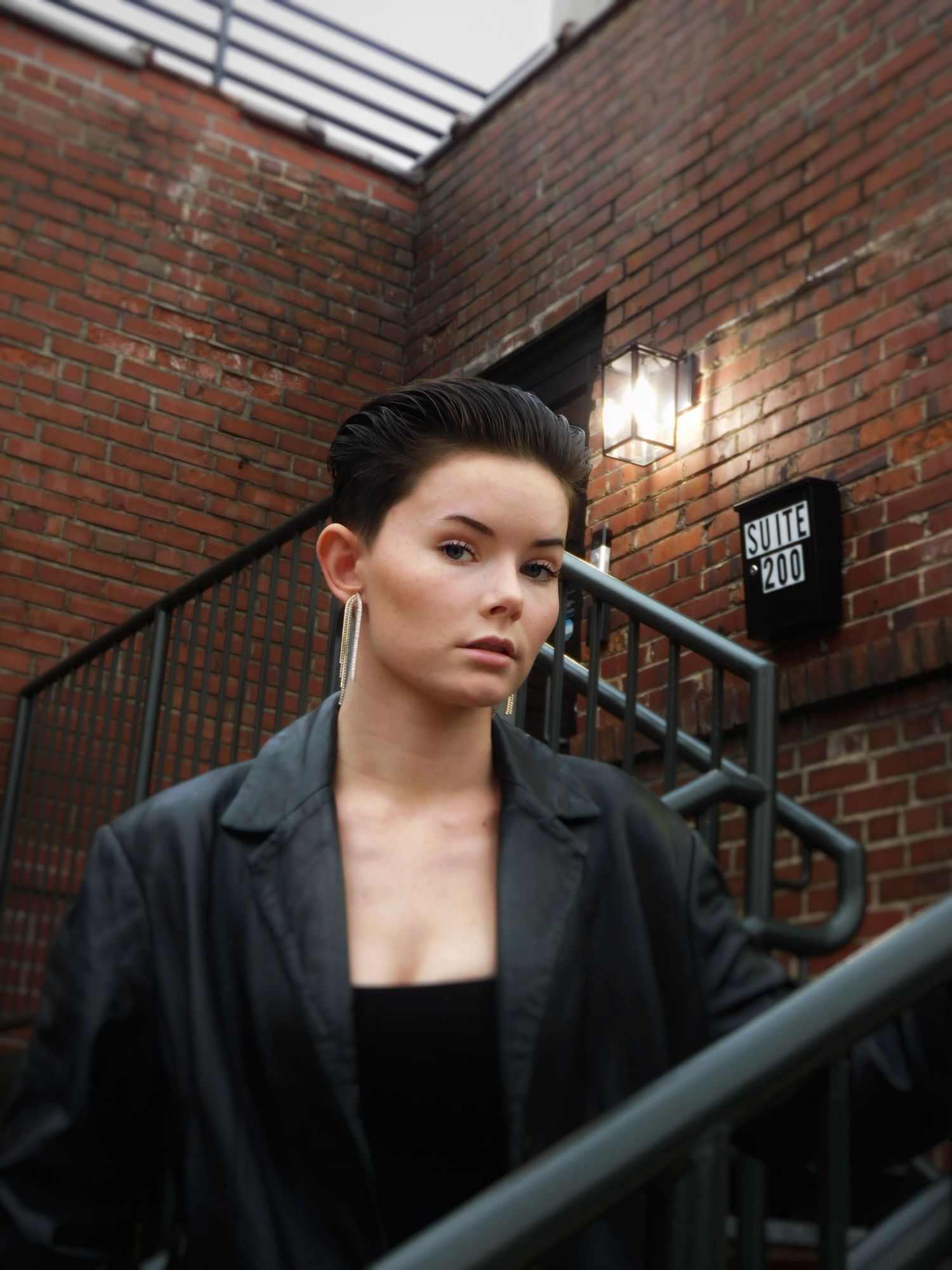 A woman with a slicked-back undercut wearing a black blazer poses on a brick building staircase.