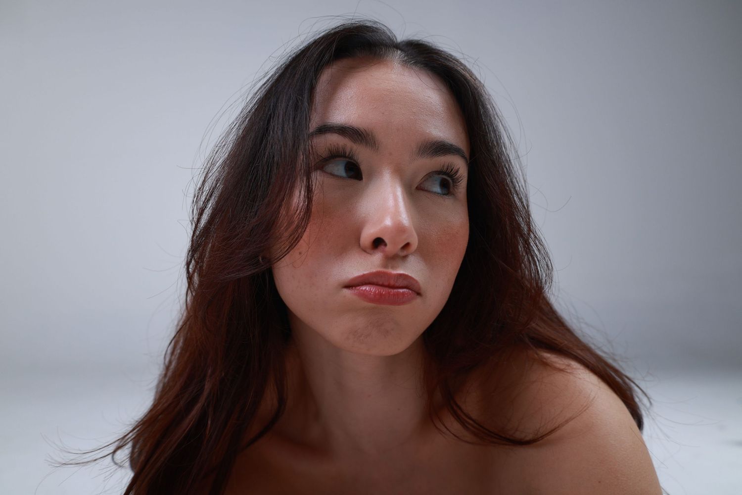 Young woman with long brown hair gazes upward thoughtfully against a light gray background.