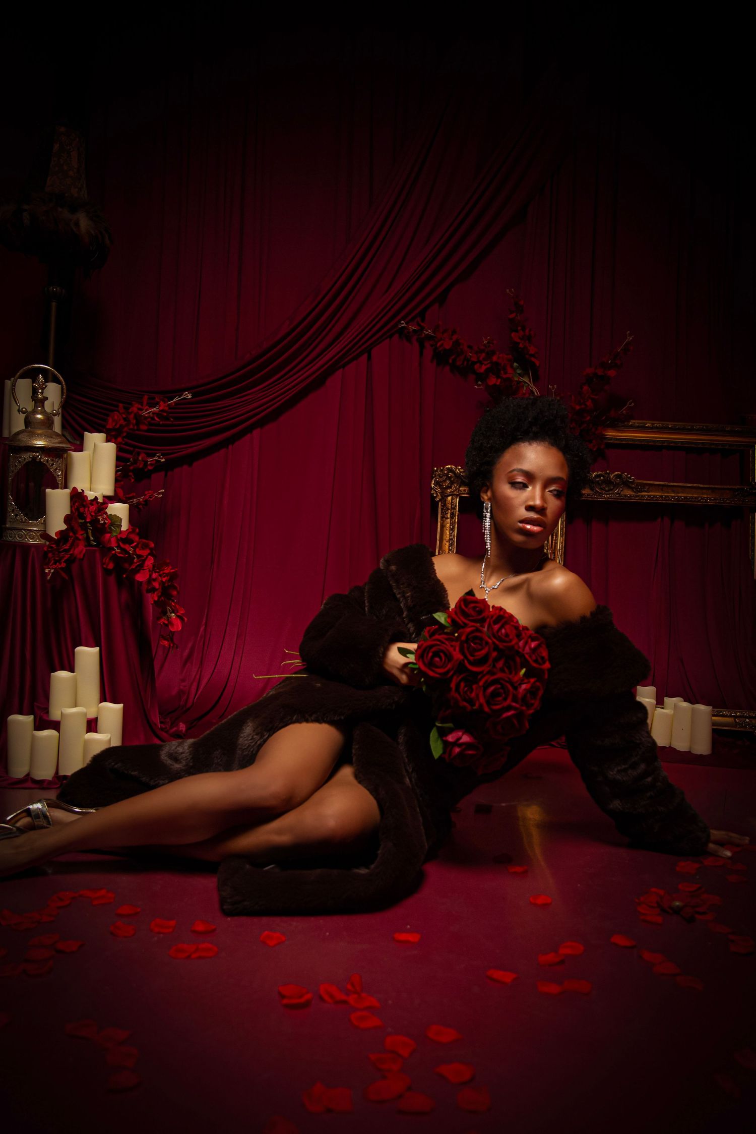 A Black woman in a black fur coat holds red roses, seated on a red floor surrounded by candles and rose petals.