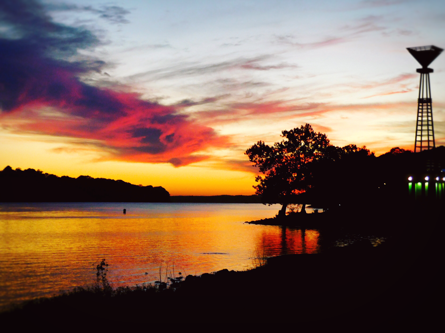 Vibrant sunset over calm lake with silhouetted trees and tower reflecting warm orange and red sky.