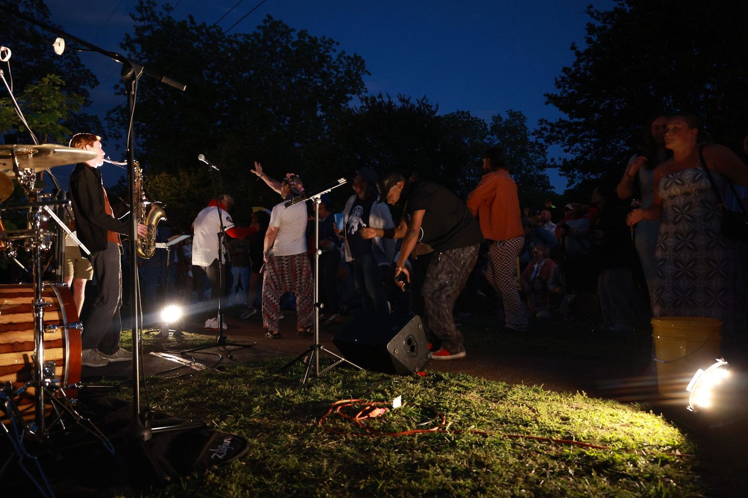 Outdoor evening concert with crowd dancing on grass near microphone stands illuminated by stage lights at dusk.