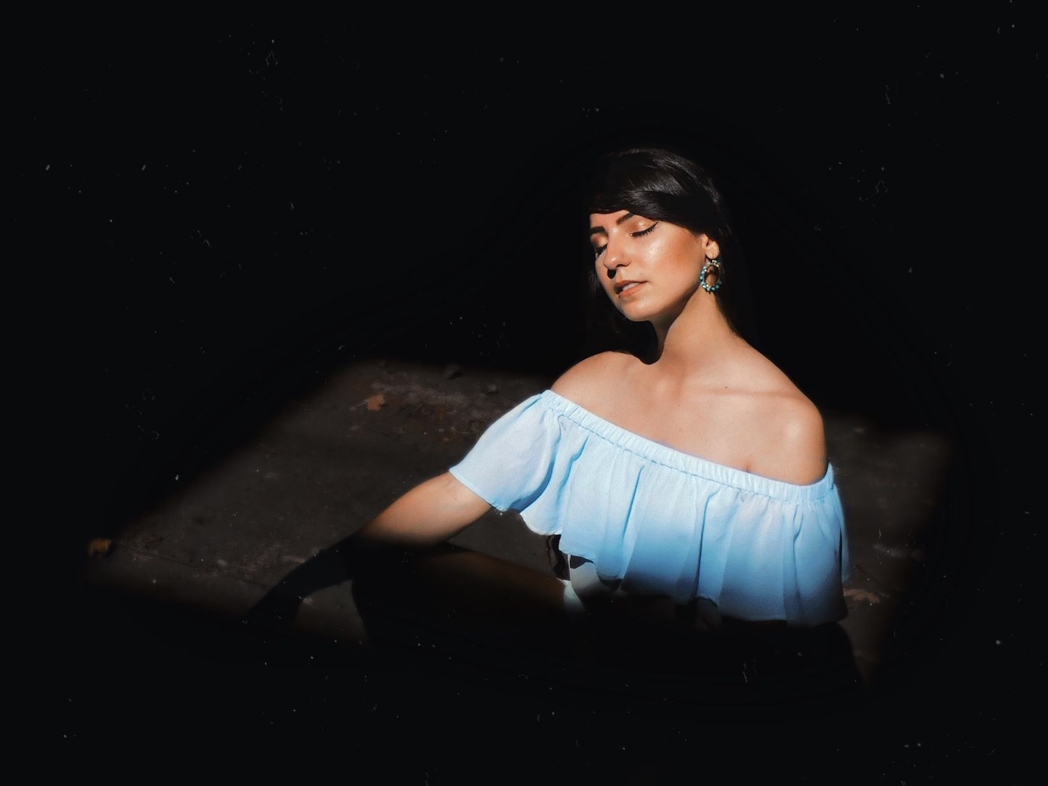 Woman in blue off-shoulder top poses gracefully against a dark background in a moody, artistic portrait.