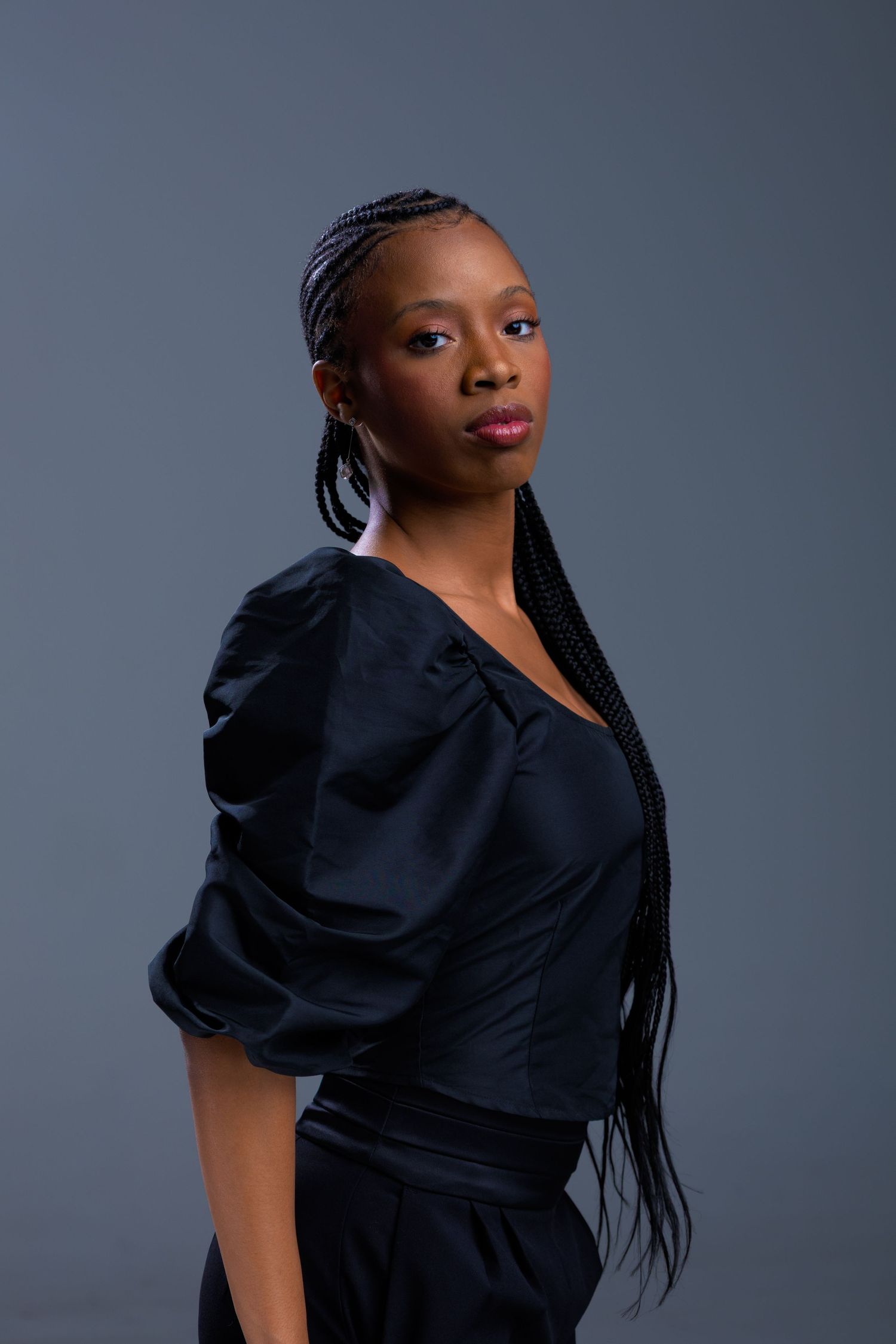 A Black woman with long box braids wearing a black puff-sleeve top poses confidently against a gray studio background.