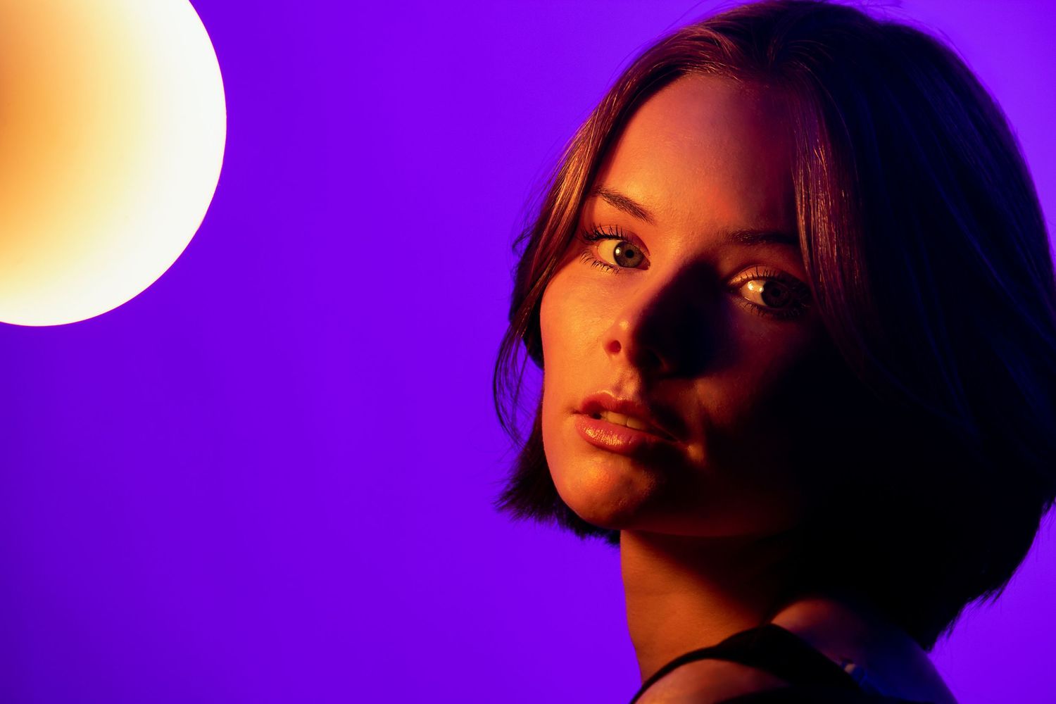 A young woman with a short bob illuminated by warm and purple neon lights in a dramatic studio portrait.