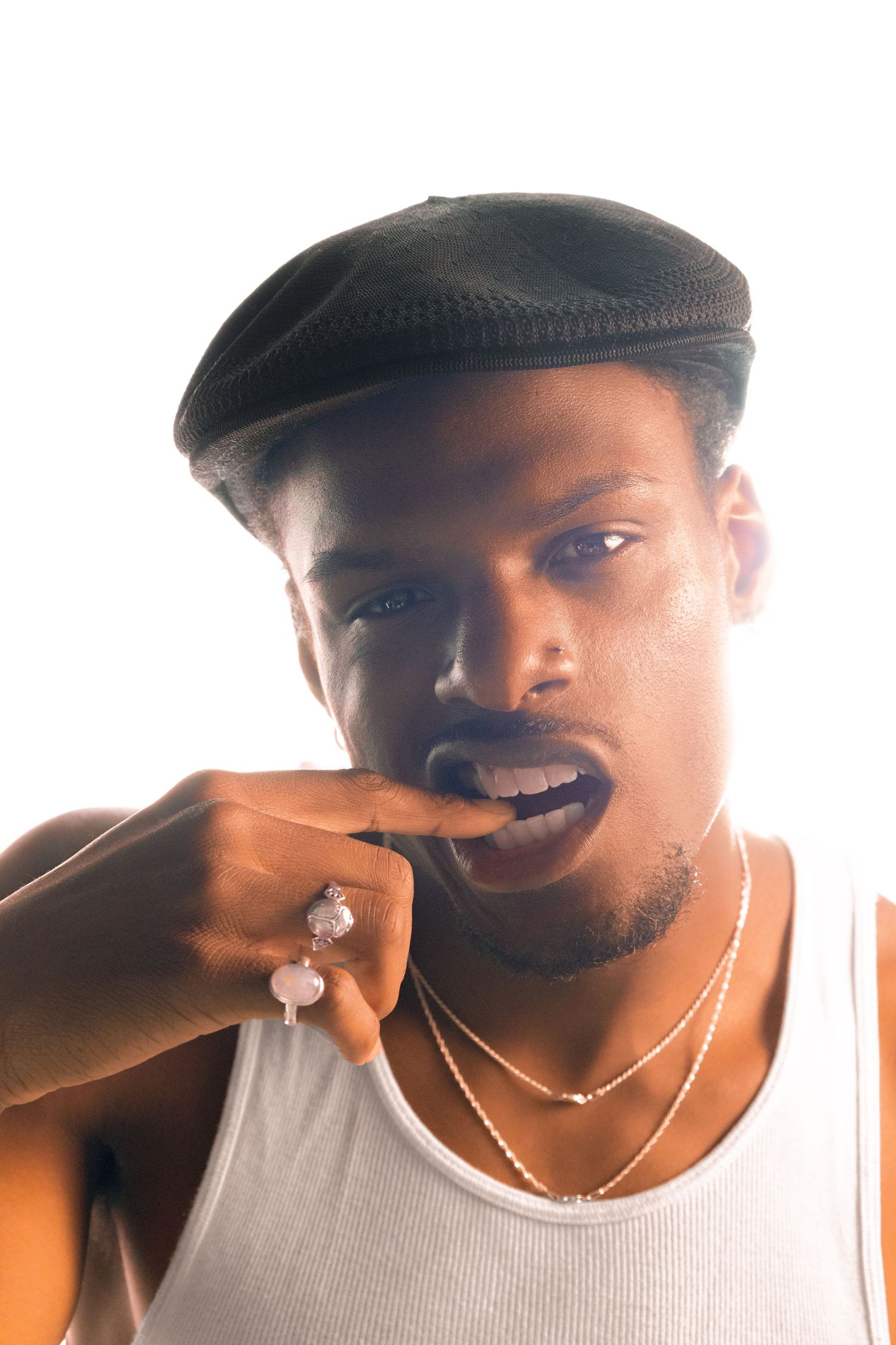 Young Black man in flat cap and white tank top wearing rings and chains, posing with finger near mouth.