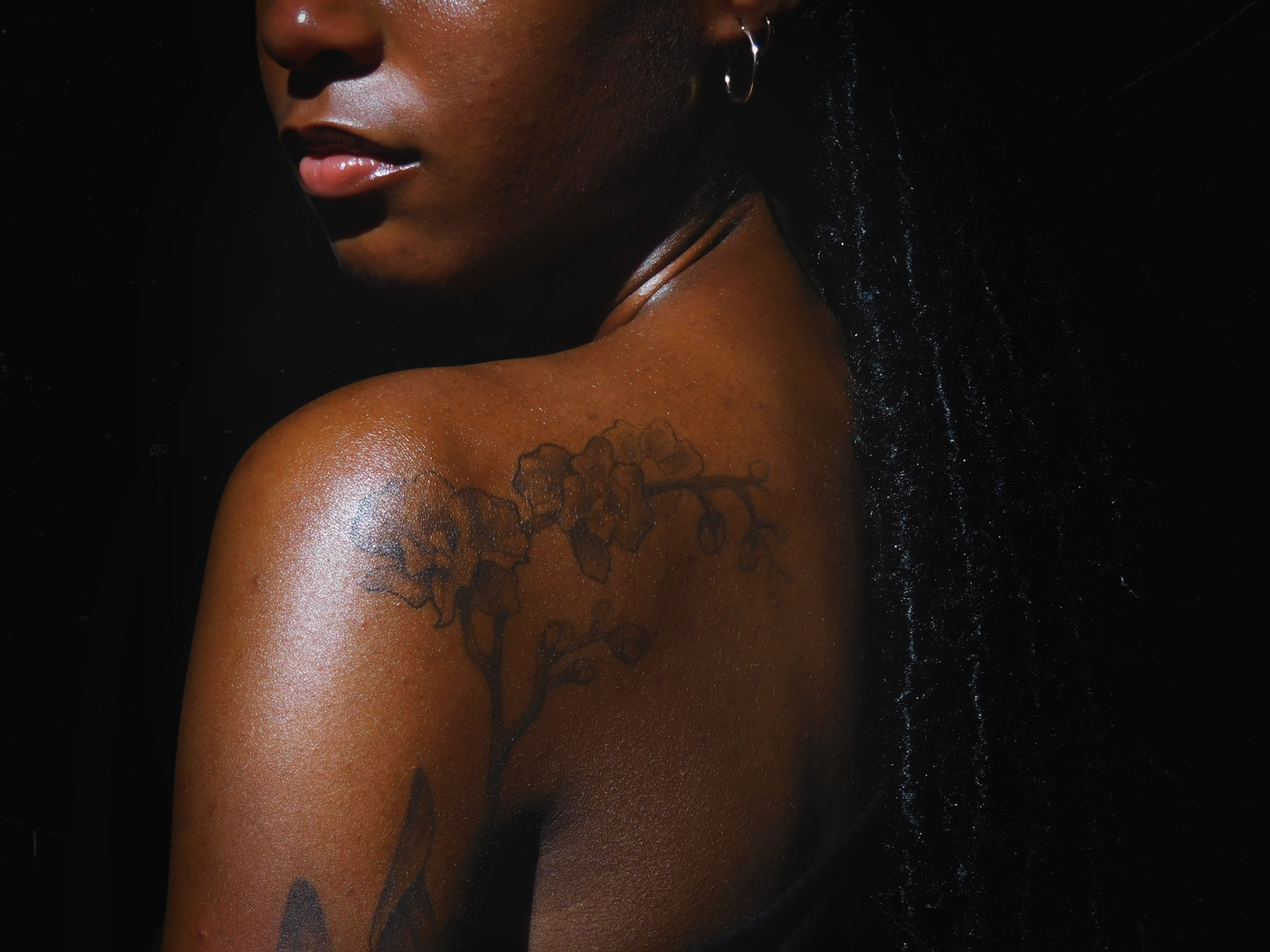 Close-up portrait showing a delicate floral tattoo on dark skin against a black background.