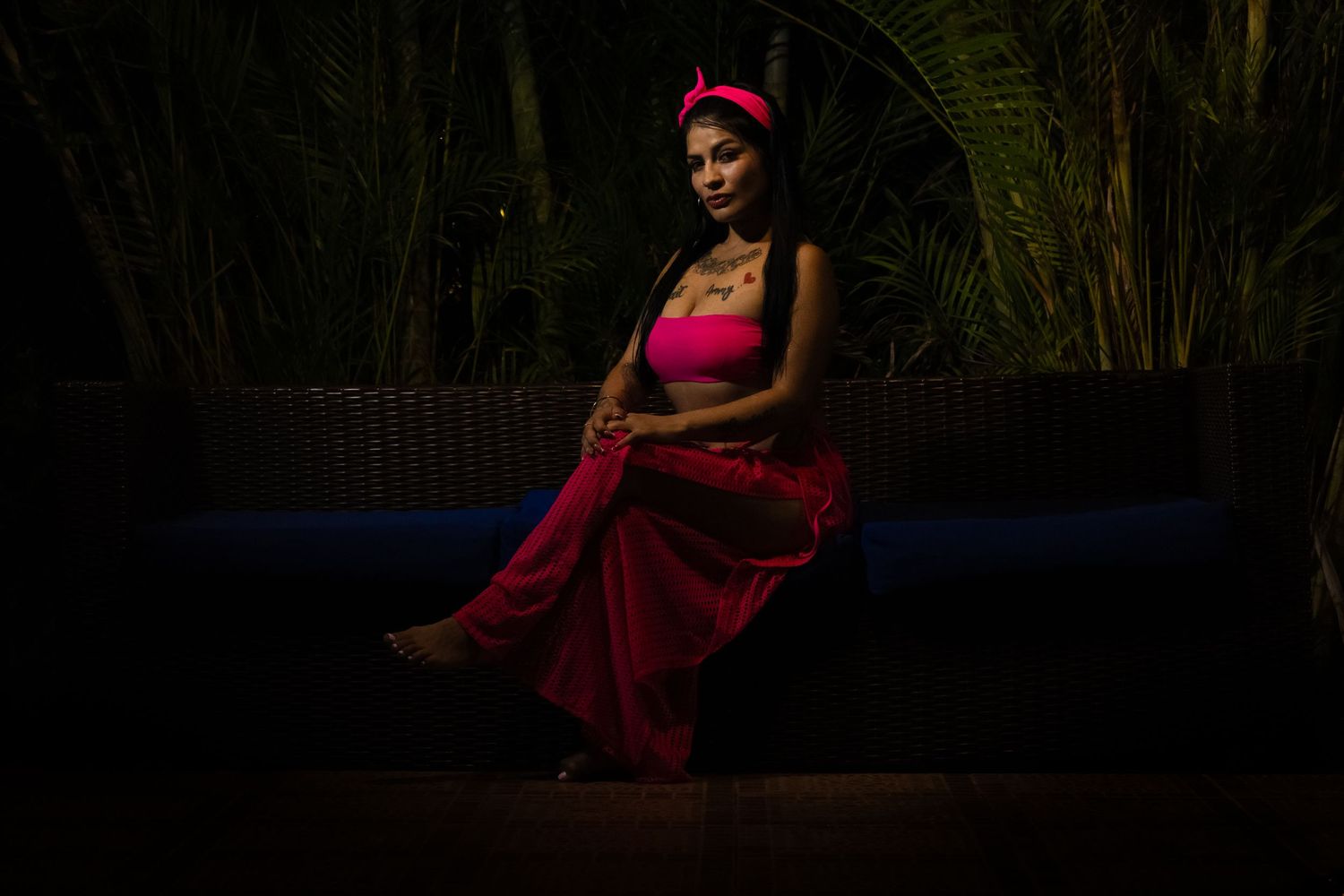Woman in pink outfit sitting gracefully on dark sofa surrounded by tropical plants in moody evening lighting.