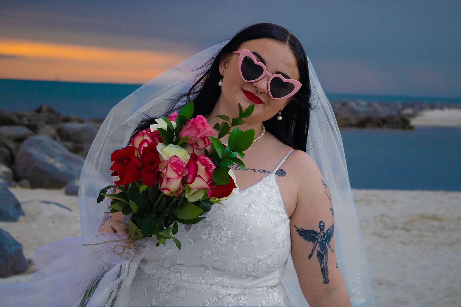 Bride in sunglasses and white dress holds red and pink roses against sunset beach backdrop on her wedding day.