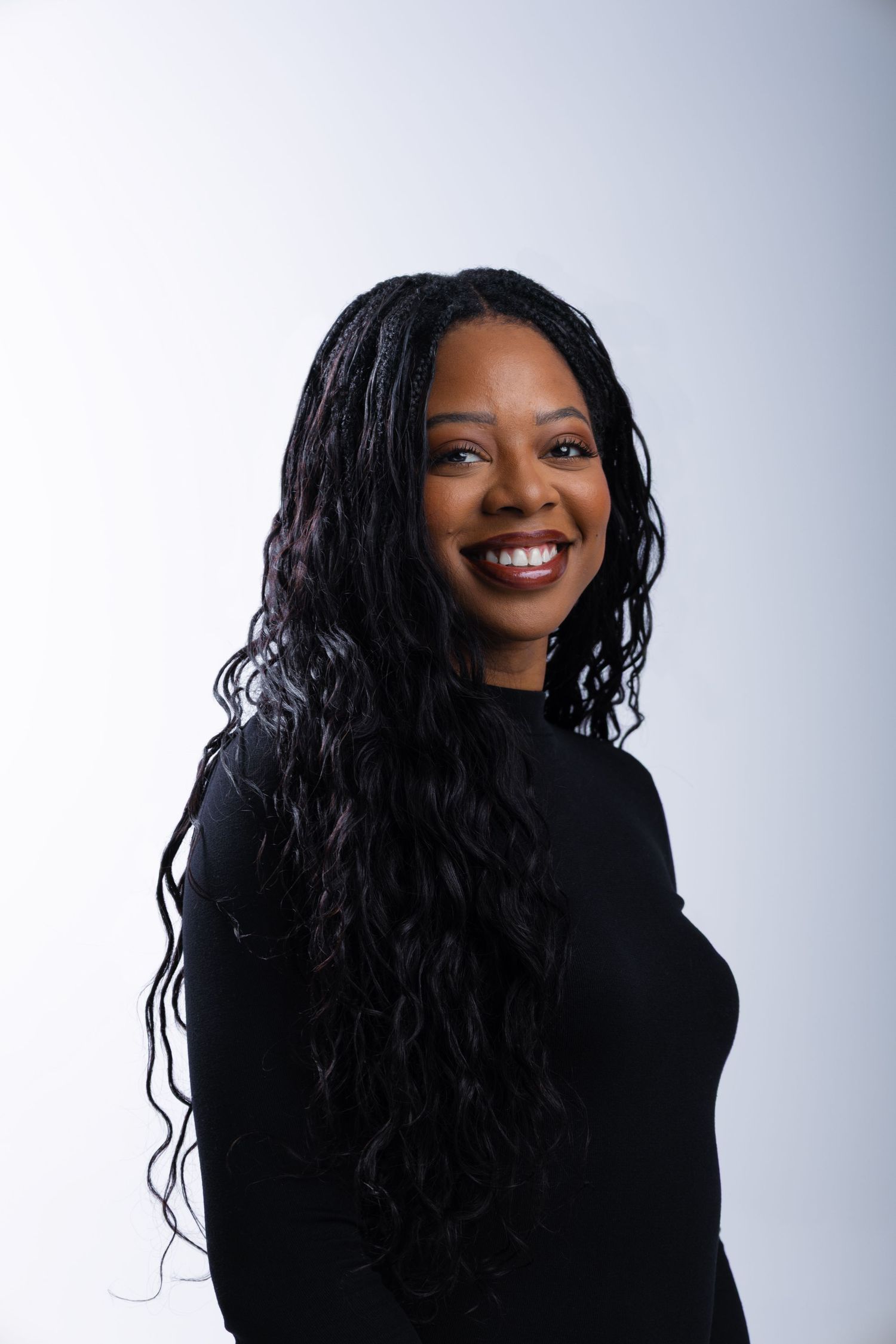Smiling Black woman with long braided wavy hair wearing a black top, posing against a light gray background.