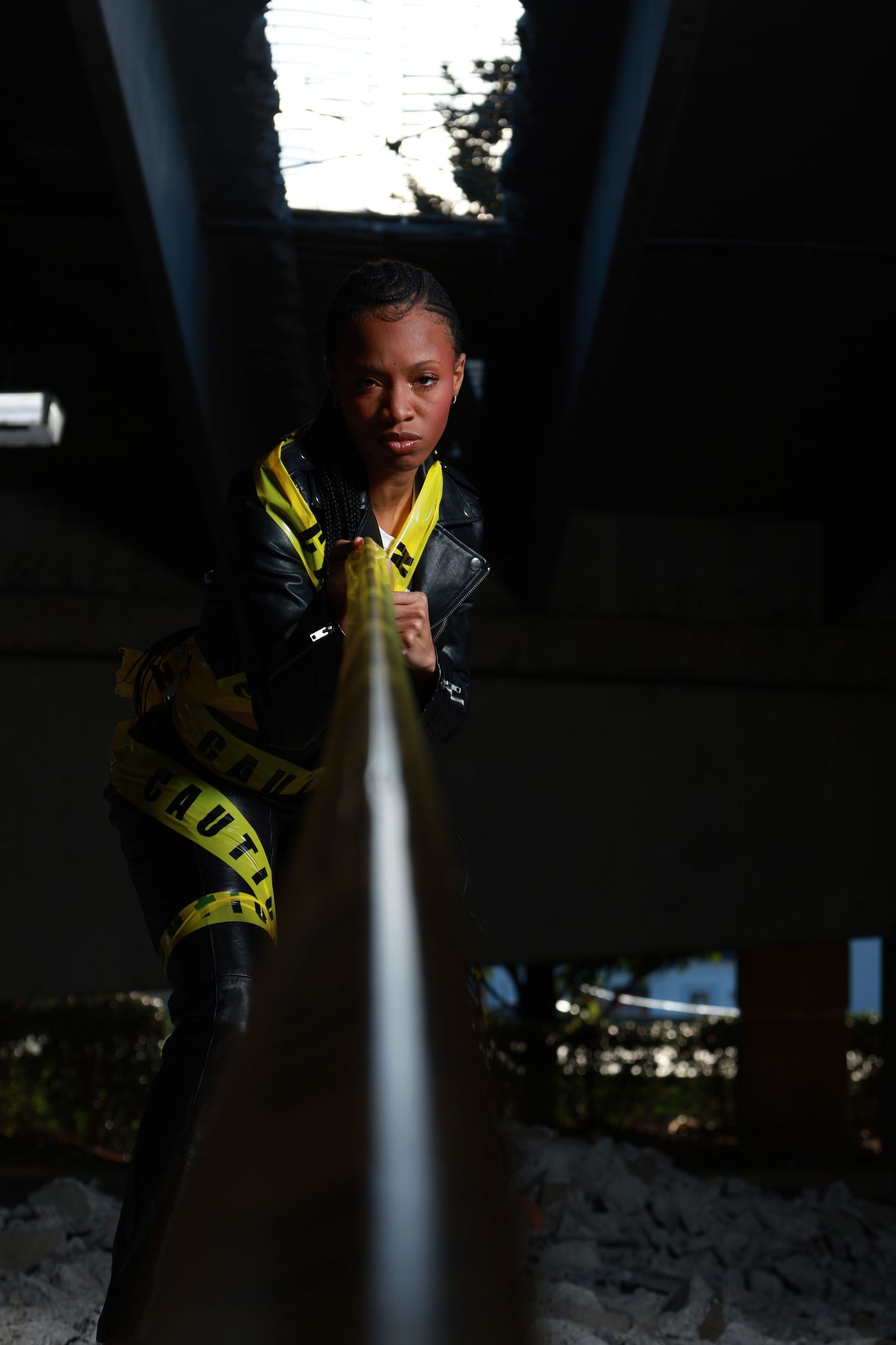 Young woman in black leather jacket and yellow caution tape outfit poses dramatically under a dark industrial structure.