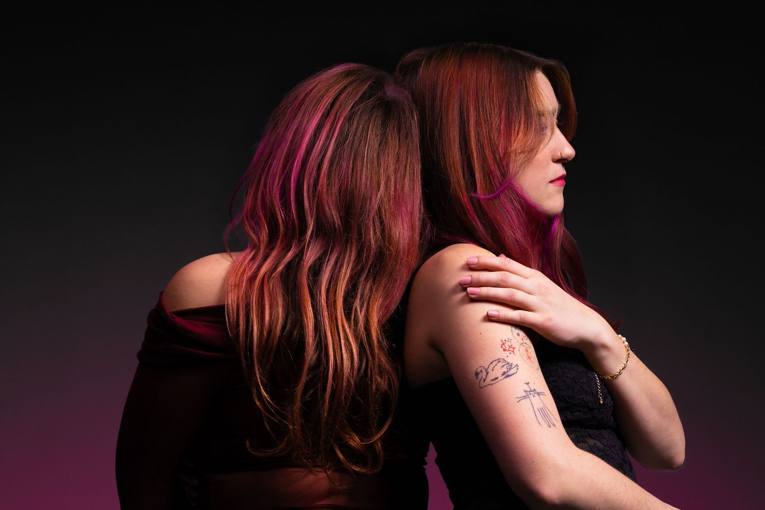 Two women with long red and pink hair stand back to back, one with a floral tattoo on her arm.