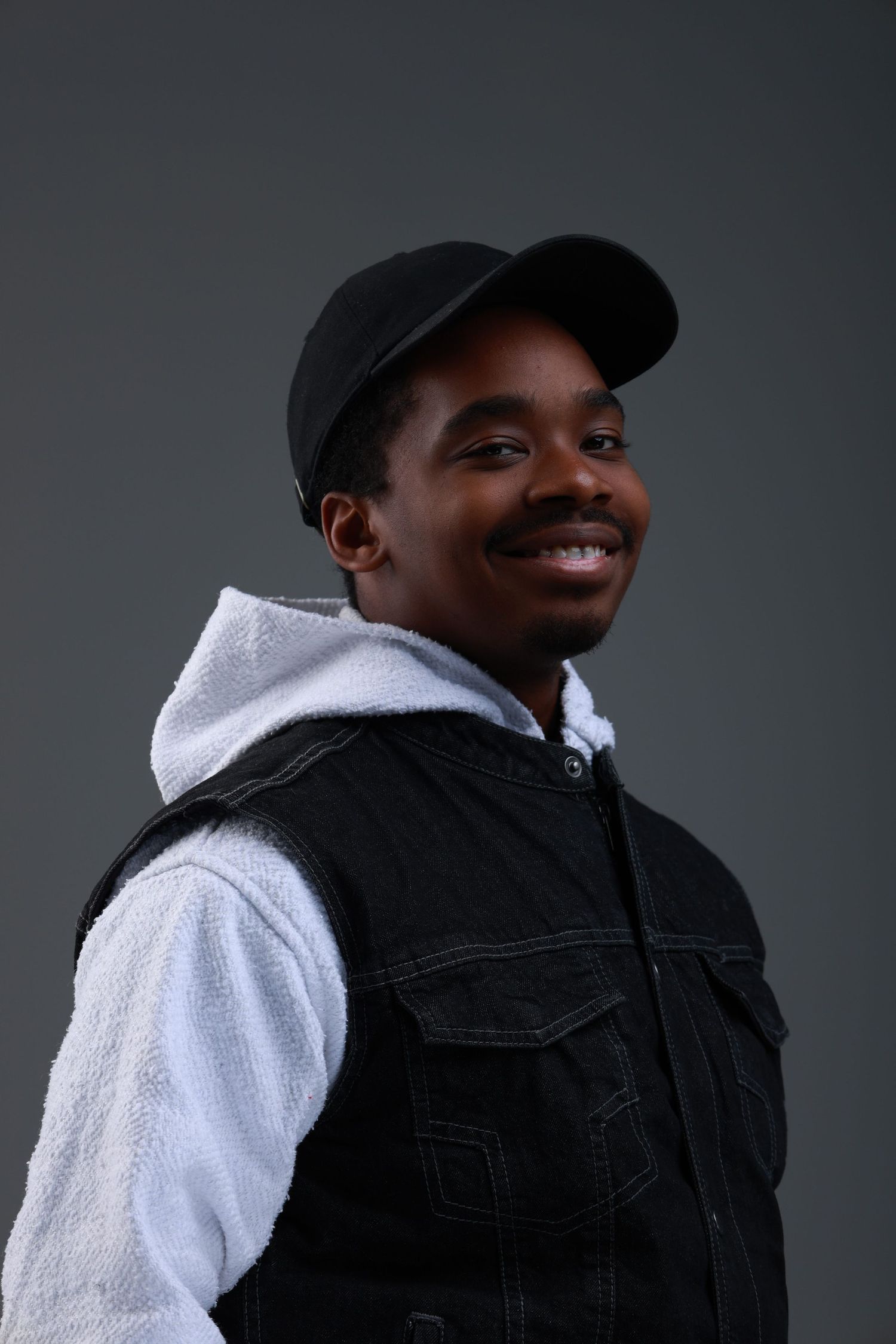 Smiling young Black man wearing a black cap and denim vest over a white hoodie, posing against a gray background.