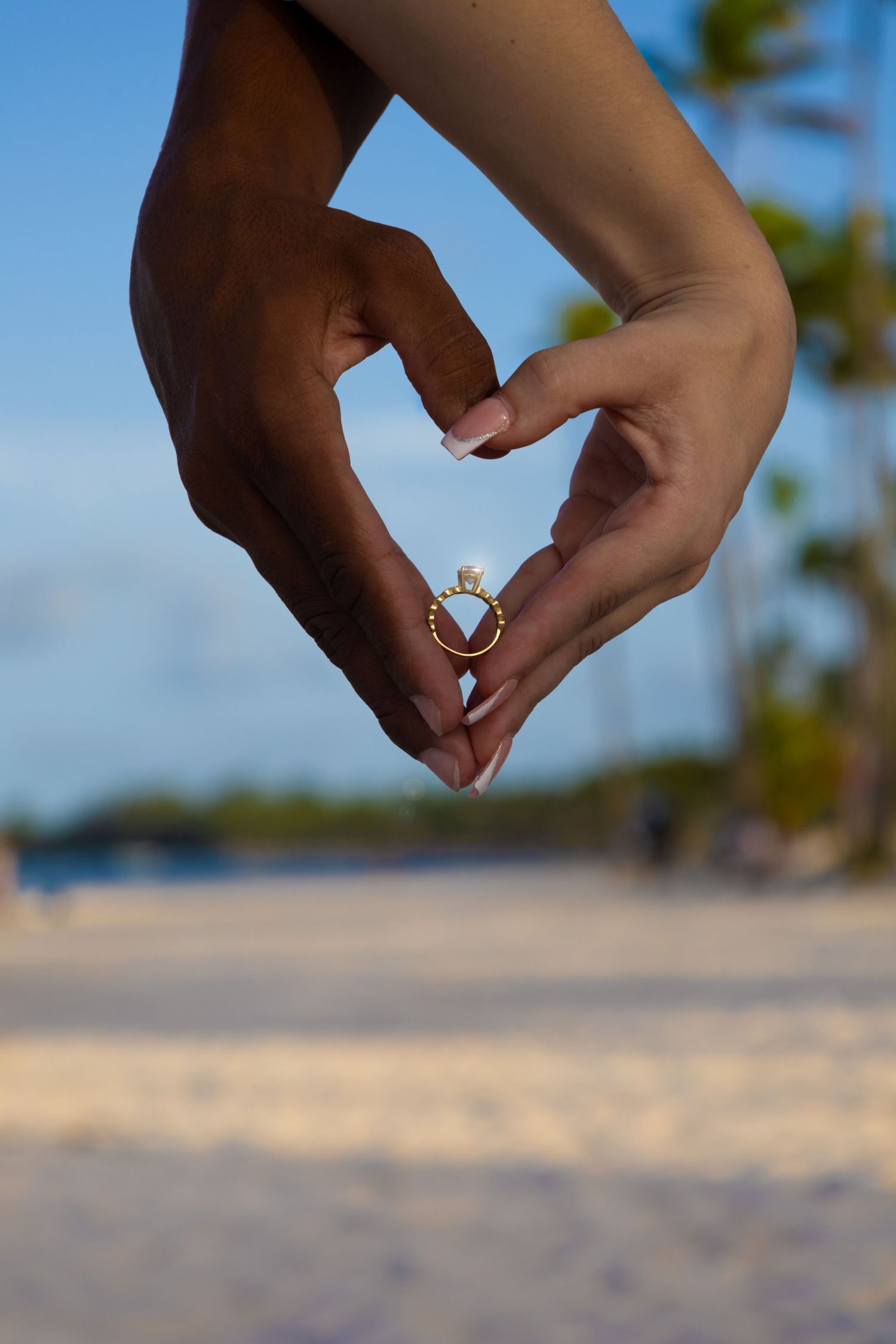 Two hands forming a heart shape around a diamond engagement ring on a sandy beach.
