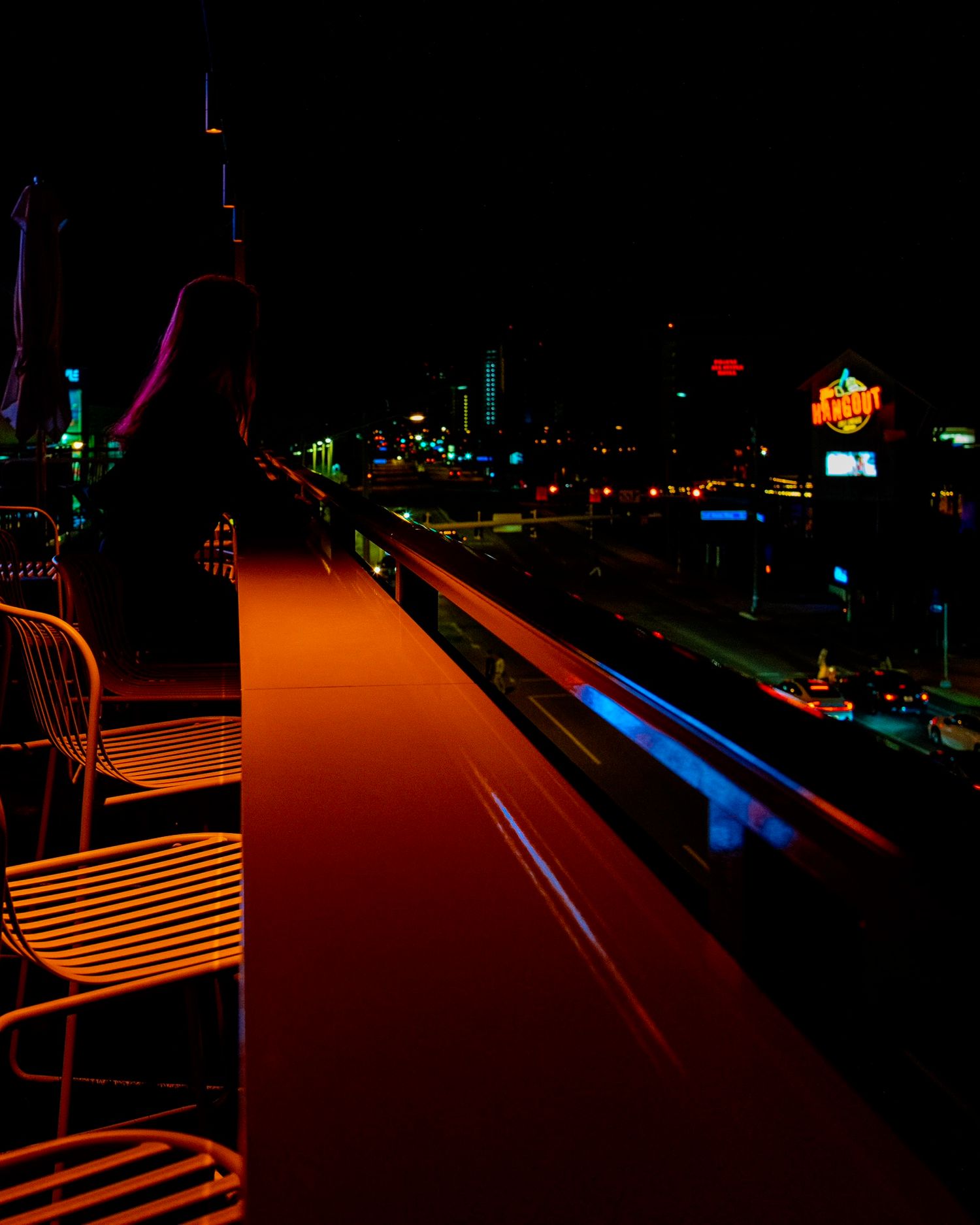 A person stands on a dimly lit rooftop bar at night with city lights and neon signs glowing in the background.