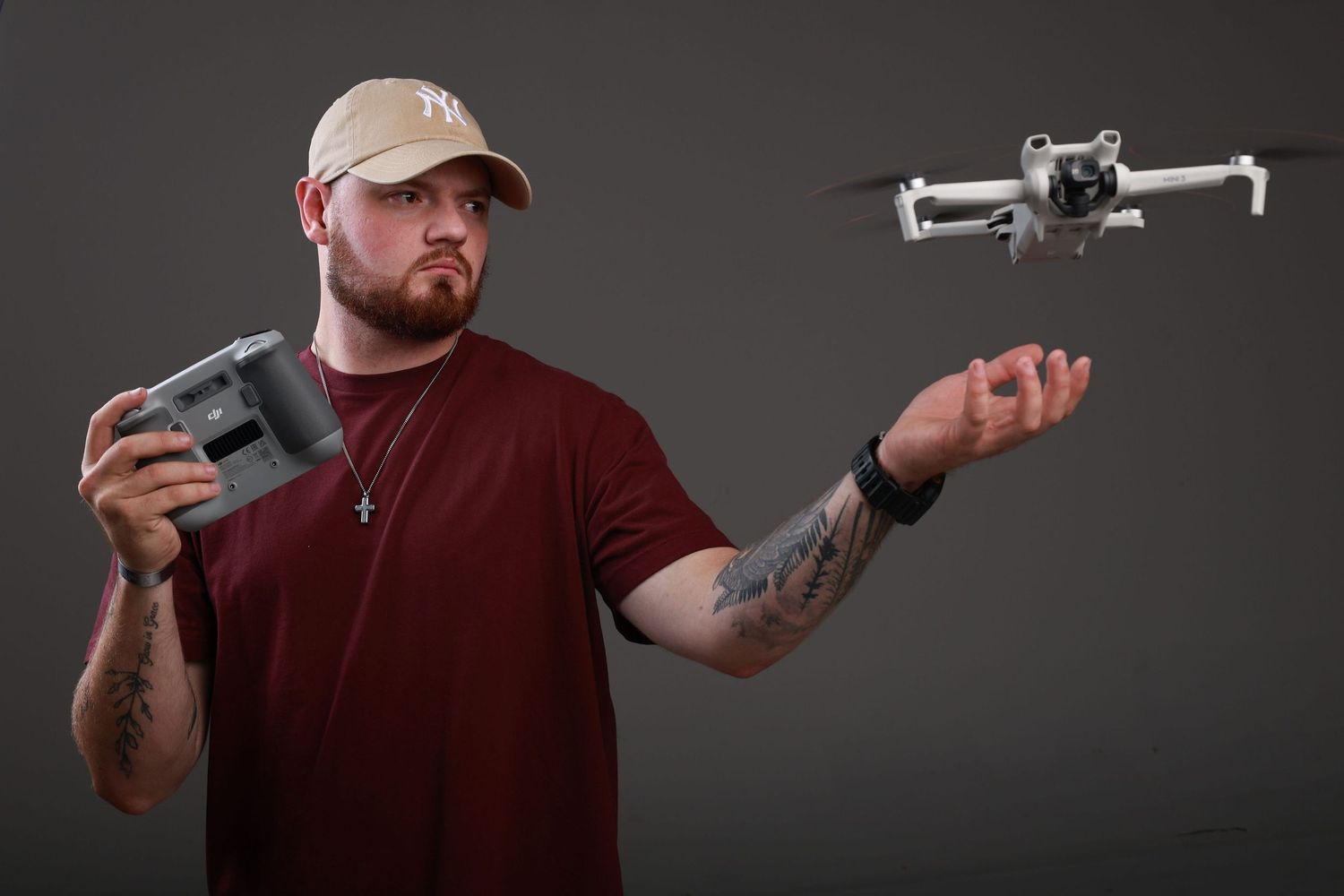 Tattooed man in red shirt and tan cap holds drone controller while balancing a small DJI drone on his outstretched hand.