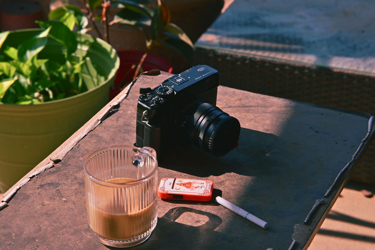 A black mirrorless camera beside an iced latte and cigarettes on a rustic outdoor table with green plants in sunlight.