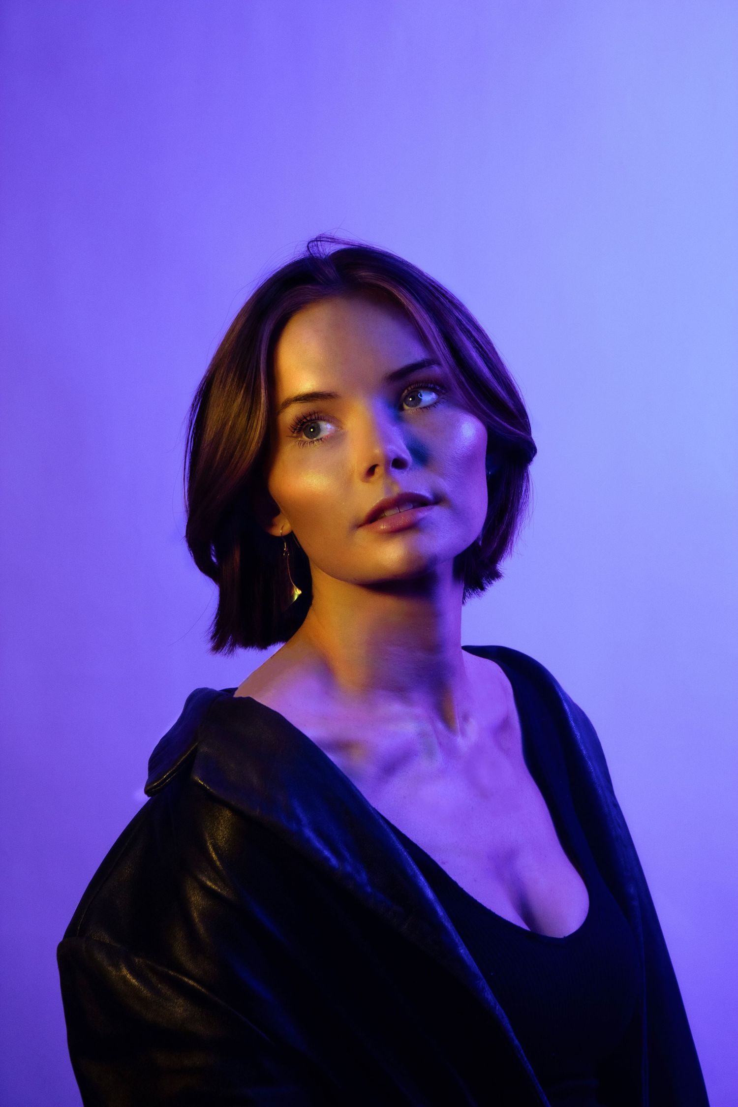 Woman in black leather jacket with short brown hair posed against purple background with colorful studio lighting.