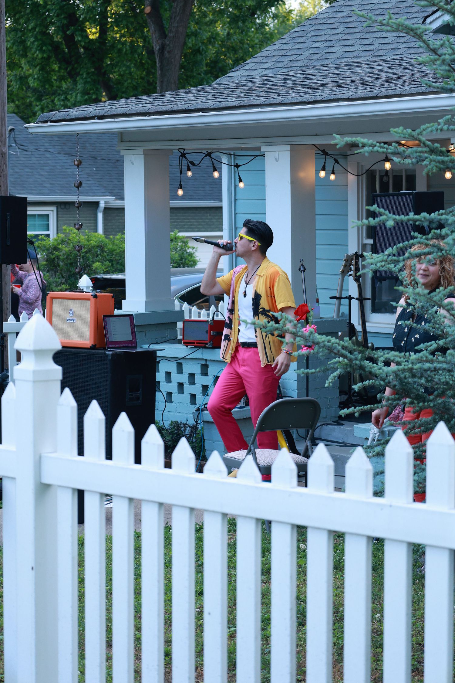 Singer in pink pants and yellow jacket performing at an outdoor backyard concert near a white picket fence.