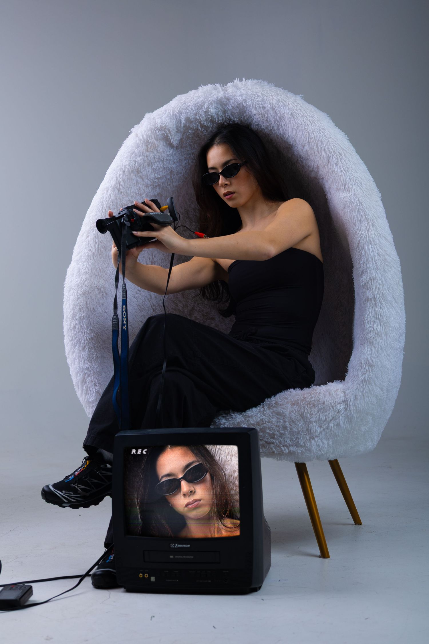 Woman in black outfit holding binoculars in fluffy egg chair, with retro TV displaying her image on the floor.