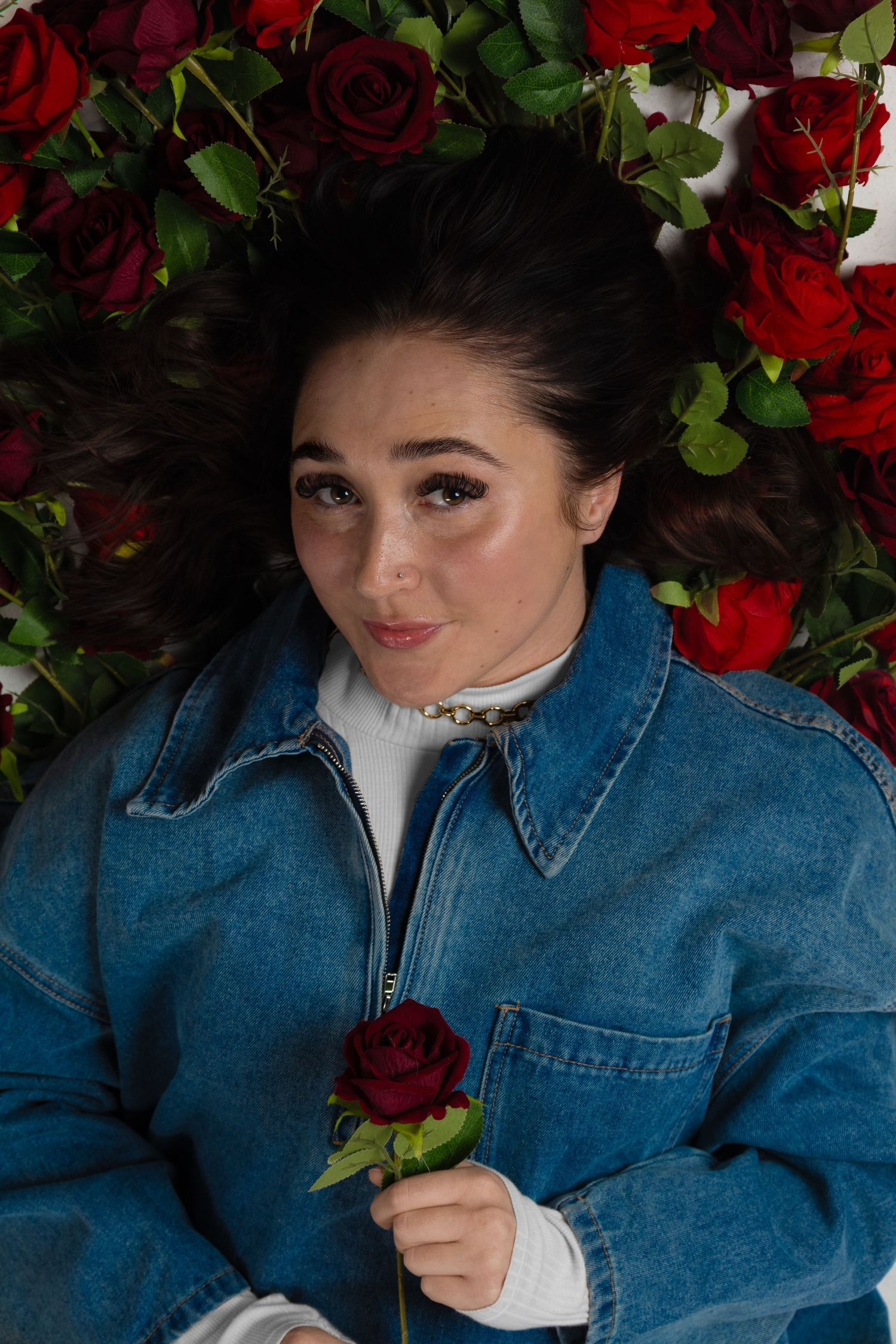 Young woman in denim jacket lying among red roses, holding a single rose, looking up at camera.
